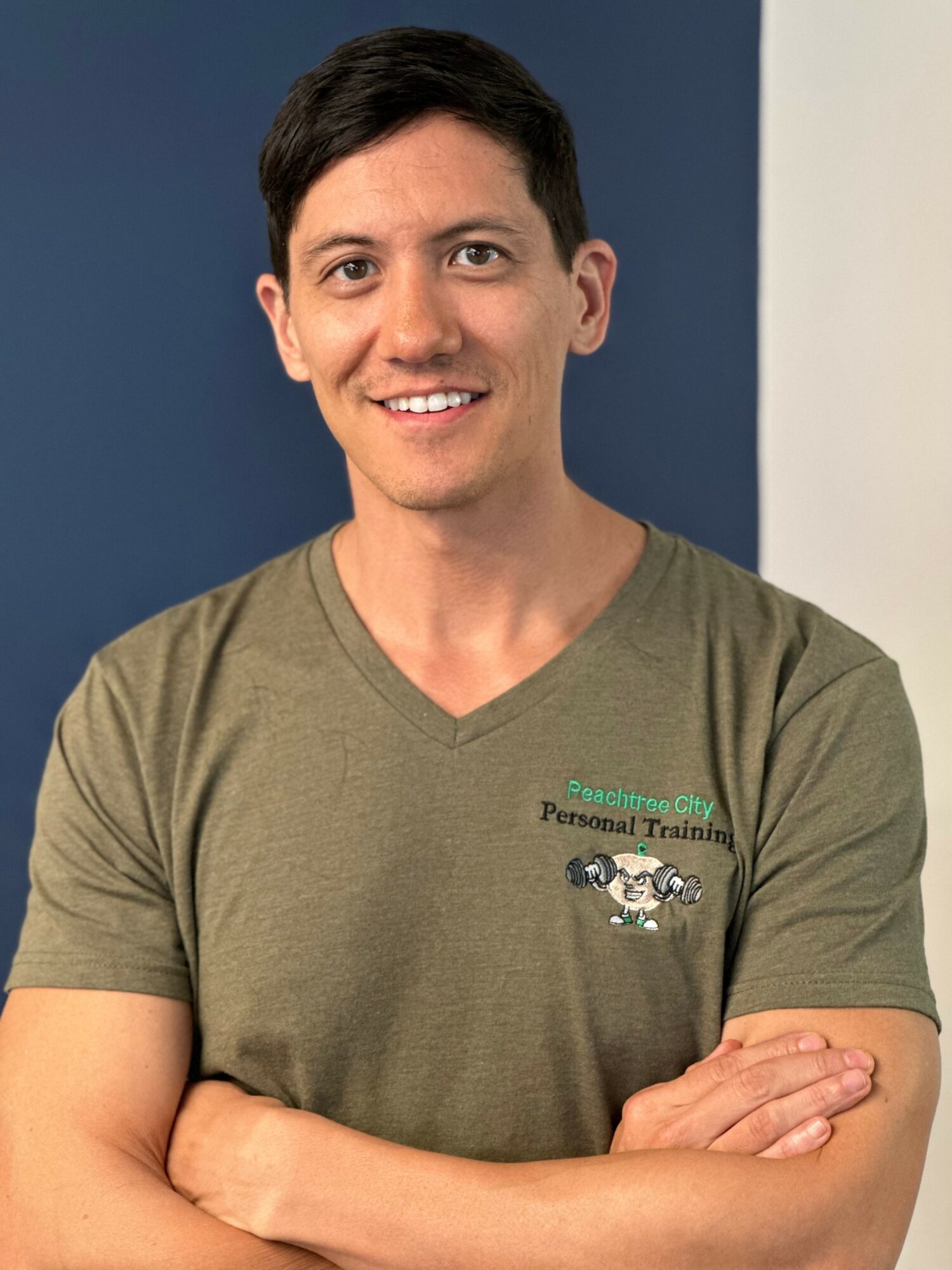 Smiling person with short dark hair wearing a green T-shirt with a logo, arms crossed, standing against a blue and white background.