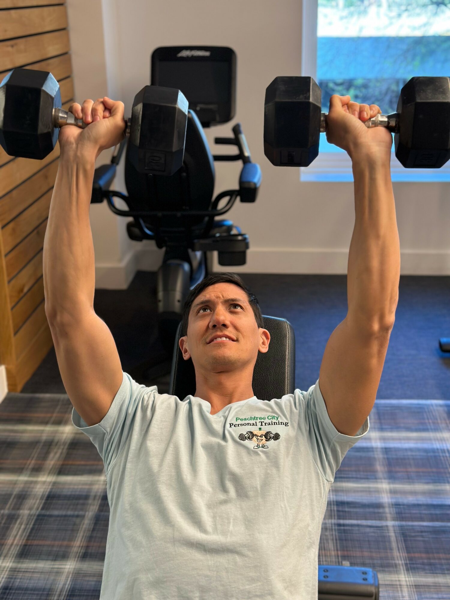 Person lying on a bench lifting dumbbells overhead in a gym, with a window and exercise equipment in background.
