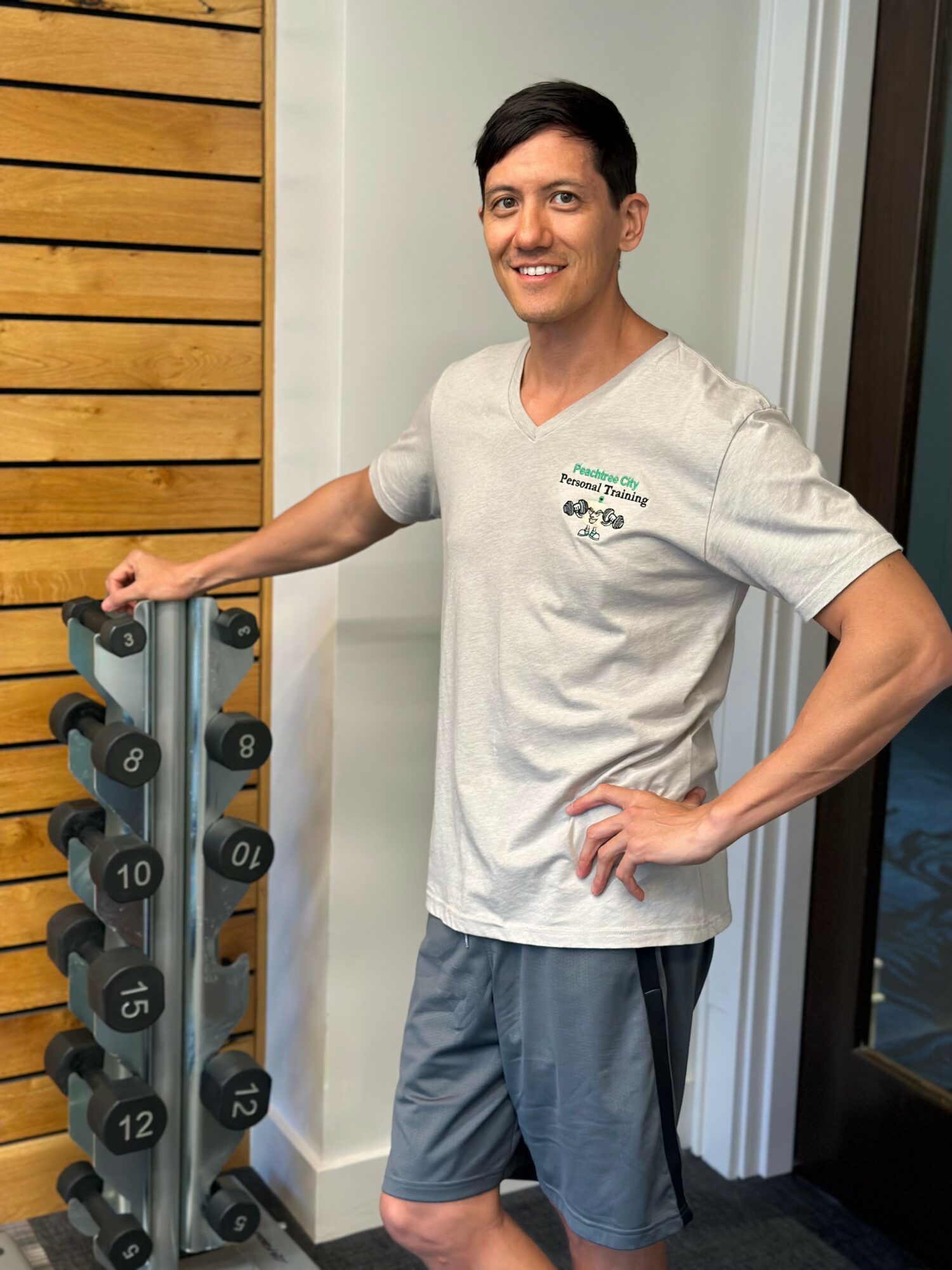 Man in gray shorts and white T-shirt standing next to a rack of dumbbells, smiling, indoors.