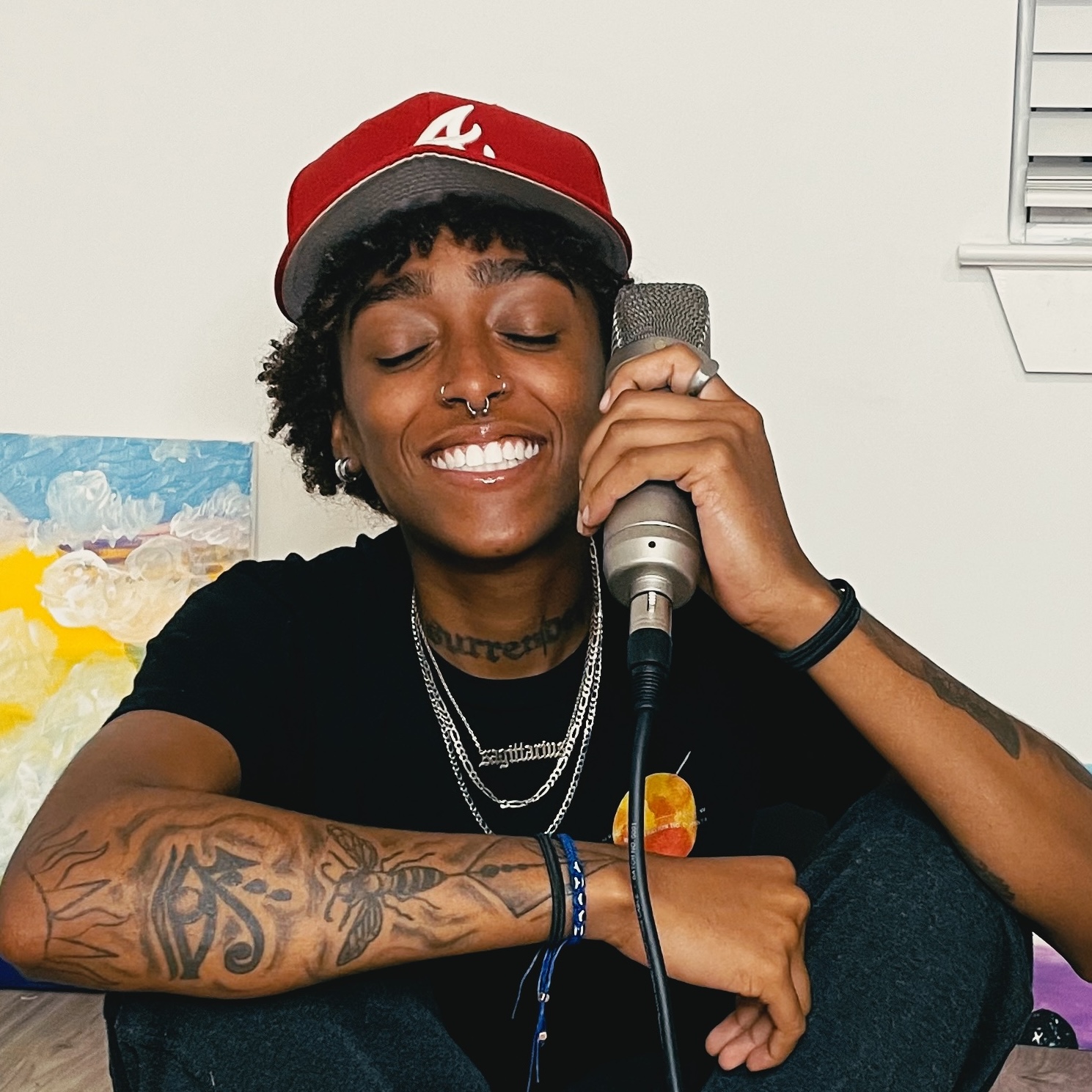 Person with short curly hair, wearing a red cap, smiling, holding a microphone, with tattoos on arm, in a room.