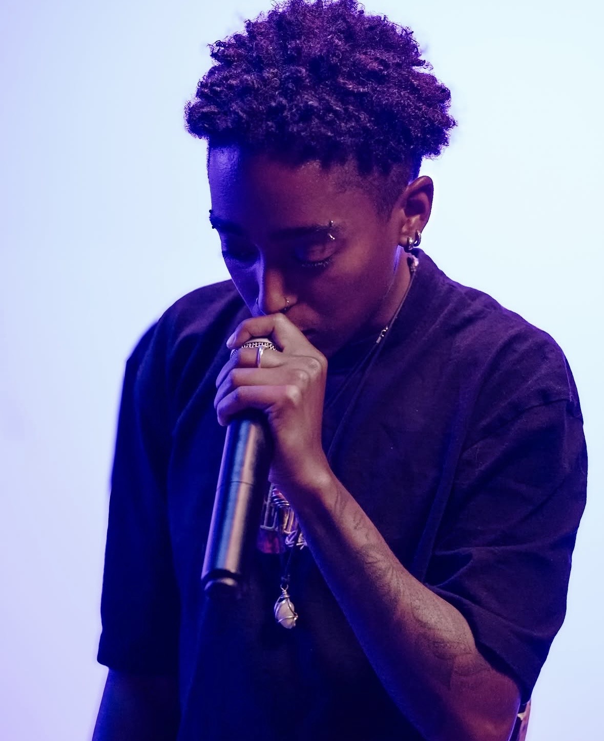Person with short curly hair holding a microphone close to face, wearing a dark shirt and jewelry, against a light background.