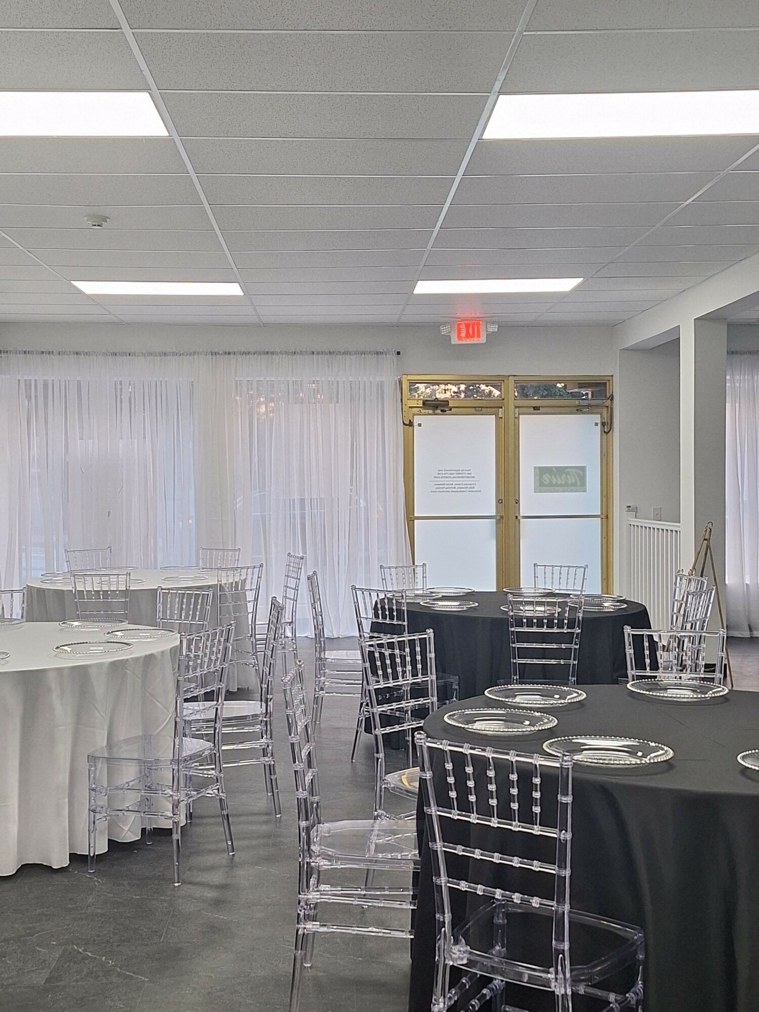 Event space with tables, chairs, and a glass door, decorated with white curtains and table settings.