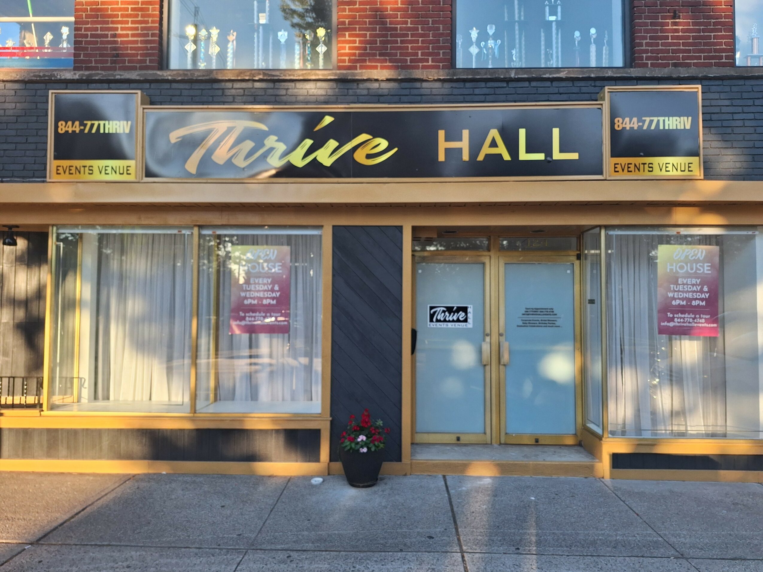 Storefront with a sign reading 'Thrive HALL' and two posters in windows, a potted plant in front, and glass doors.