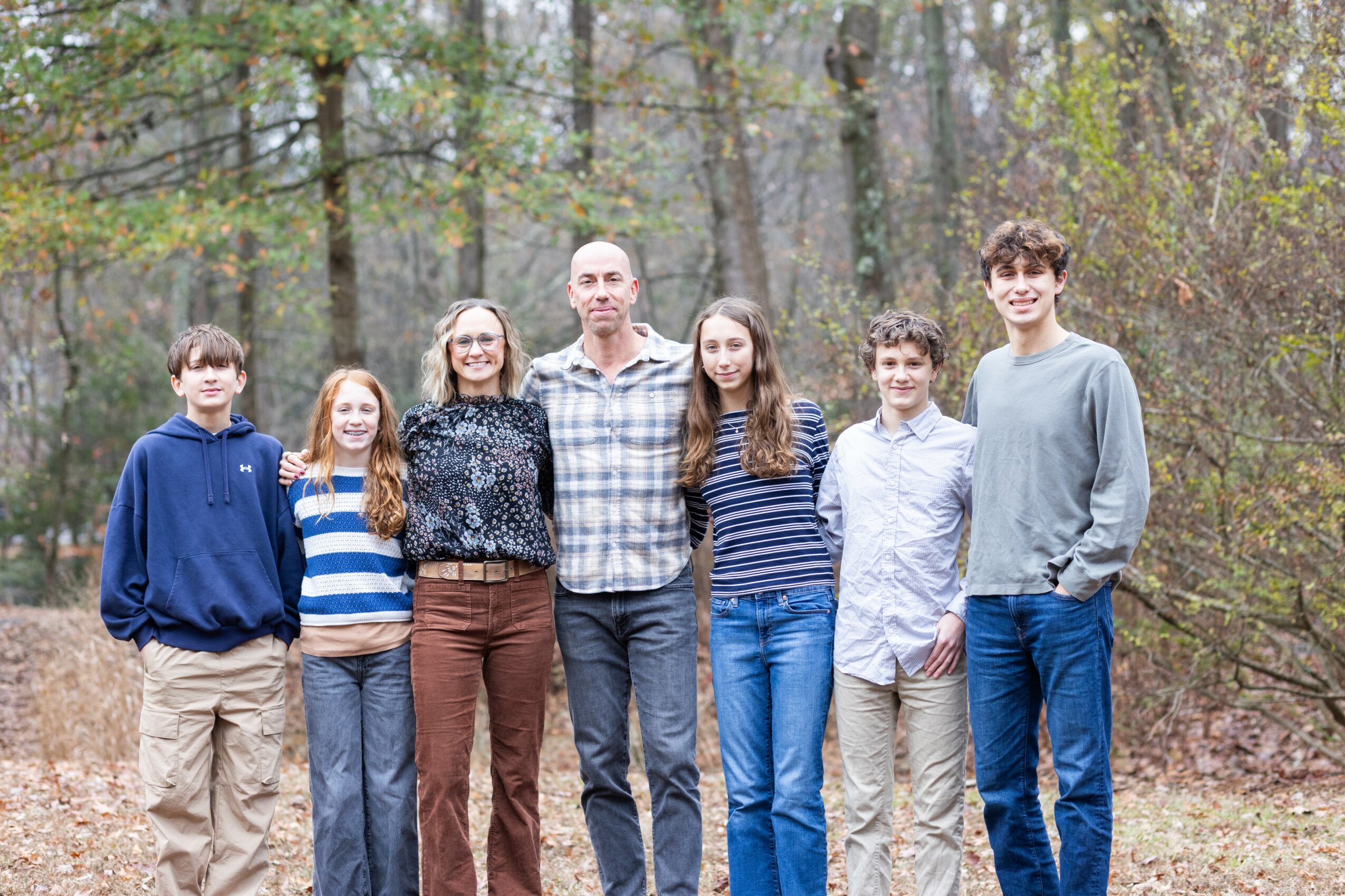 Group of seven people standing outdoors in a wooded area, posing for a photo, with trees and leaves in the background.