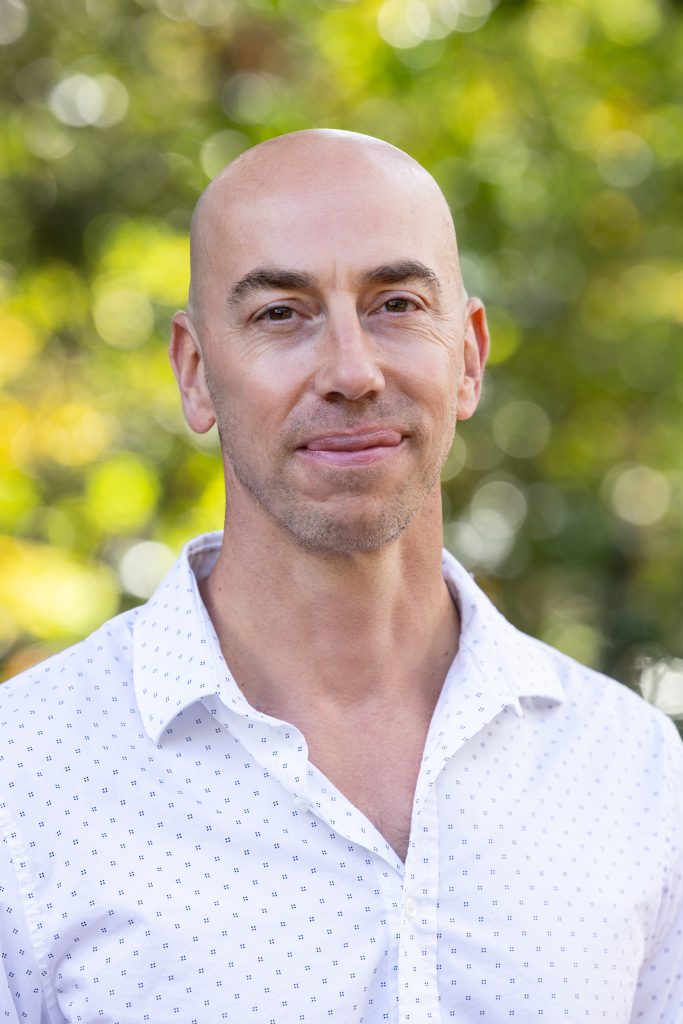 Bald man with light skin and a slight smile, wearing a white polka-dot shirt, outdoors with blurred green foliage background.