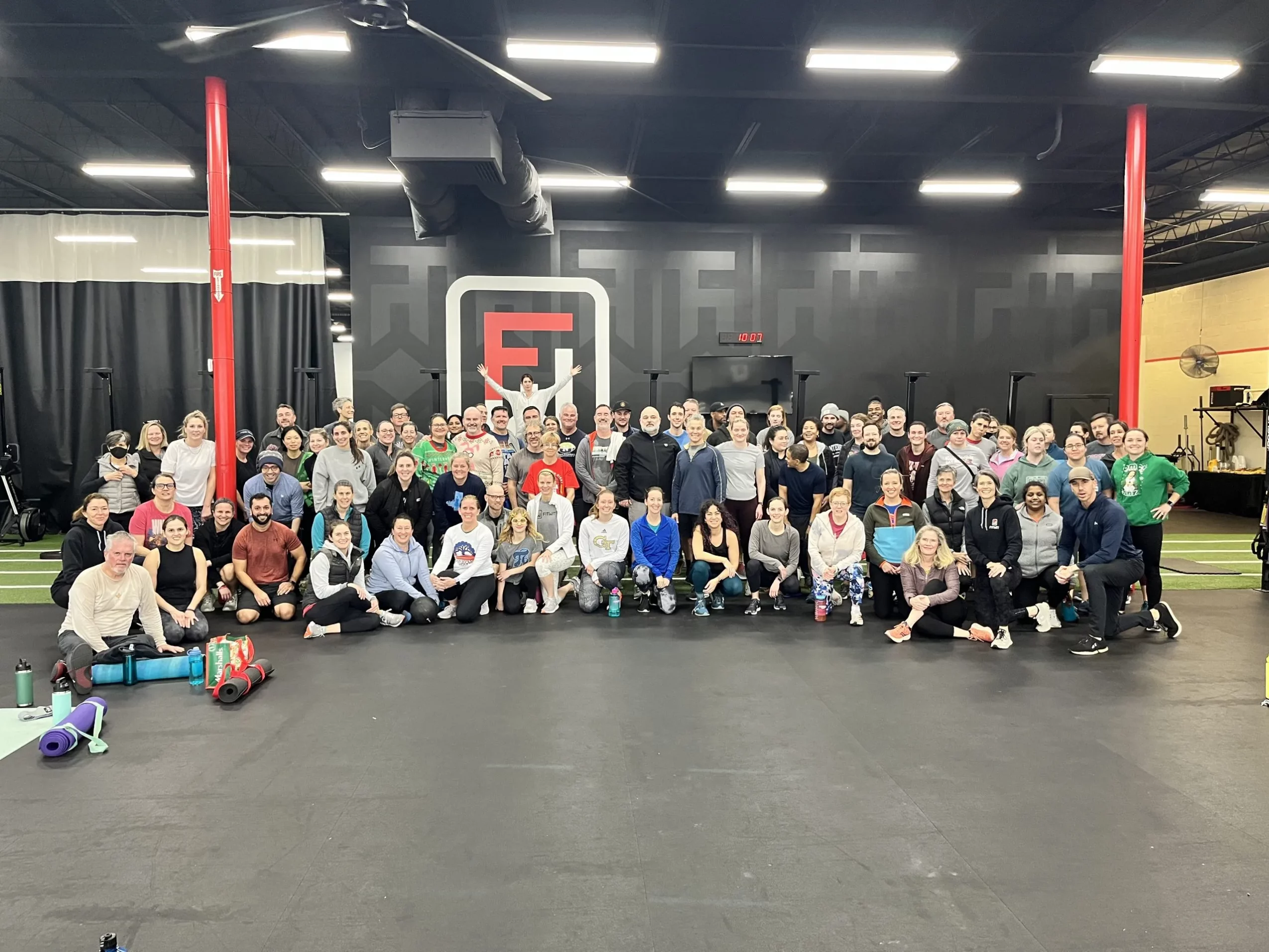 Large group of people gathered in a gym with black walls and a logo in the background.