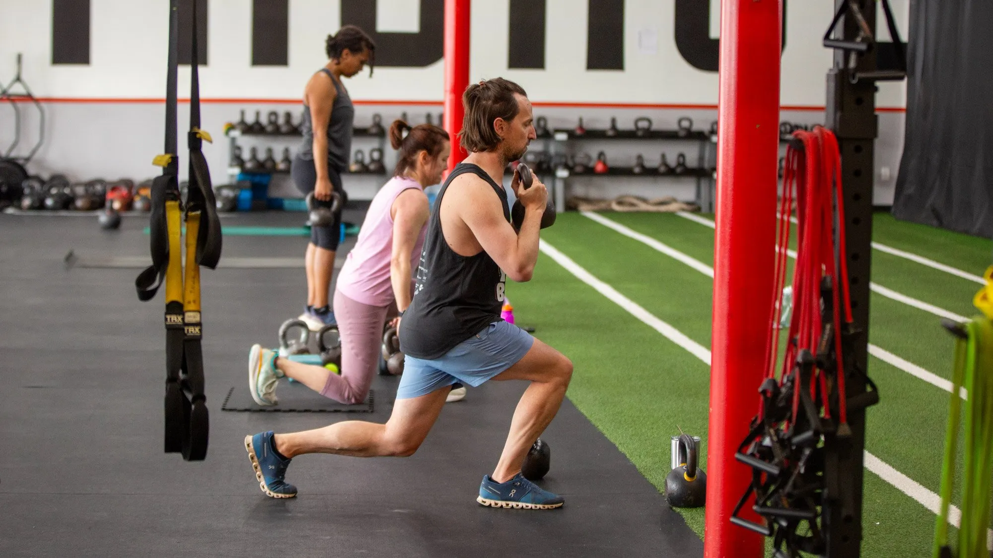 People exercising in a gym, including a man doing lunges and others stretching or working out, with gym equipment in background.