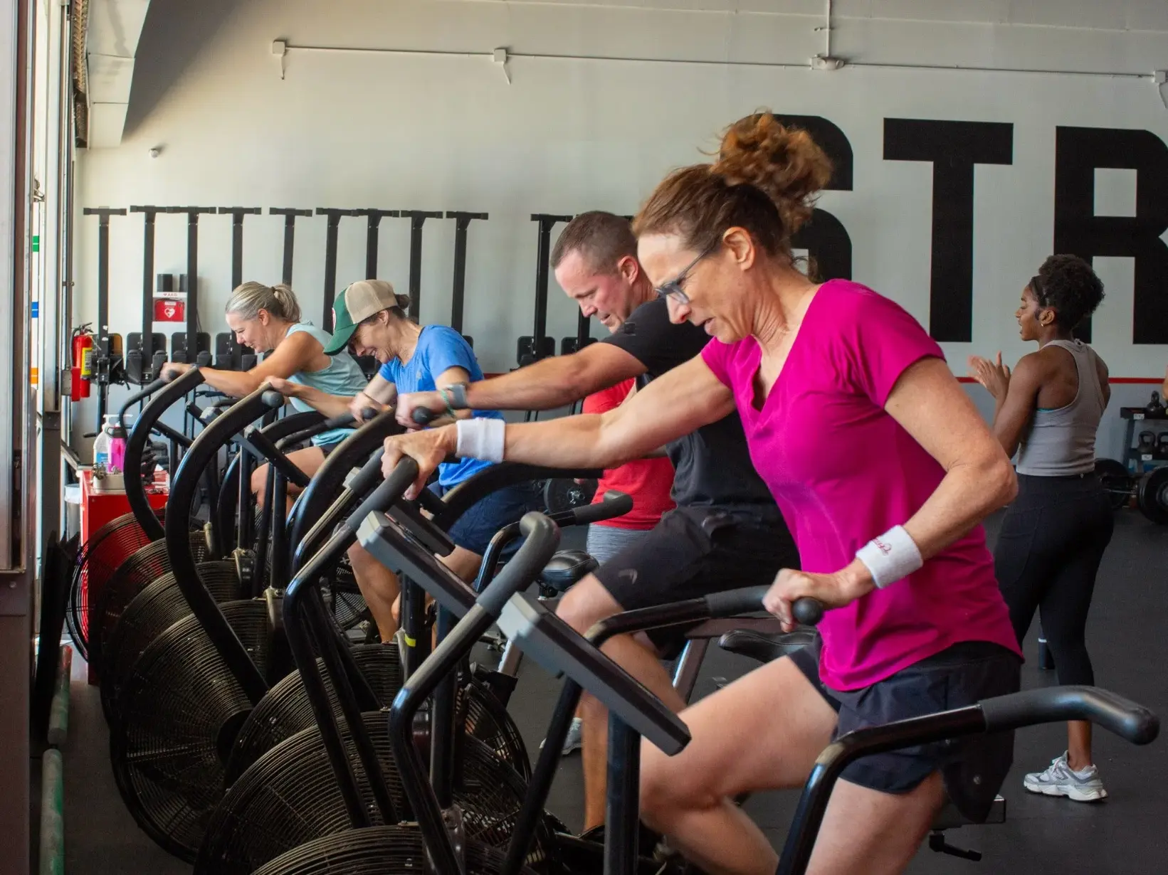 People exercising on stationary bikes in a gym, wearing workout clothes, with a railing and a wall in the background.