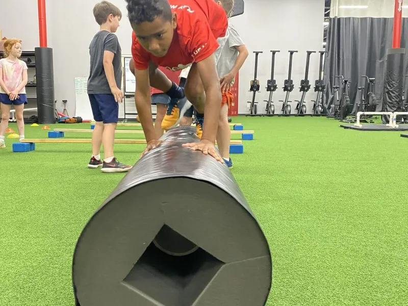 Person in red shirt leaning over a large cylindrical object in a gym with children in the background.