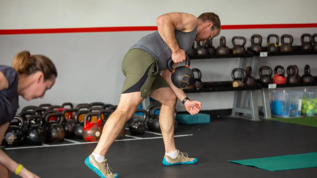 Man lifting a kettlebell with assistance from a trainer in a gym, with kettlebells on shelves in the background.