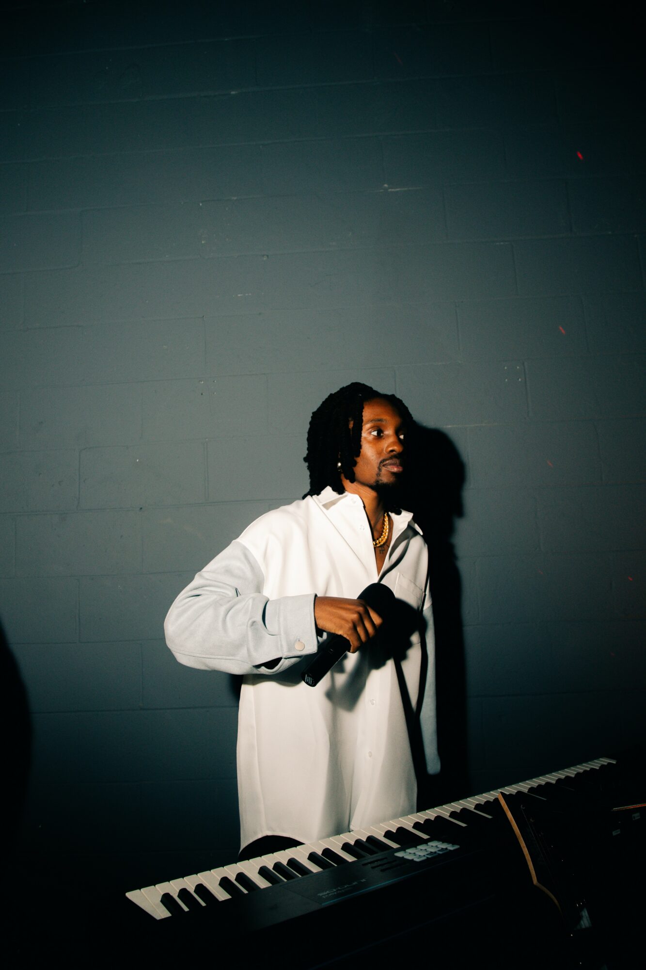 Person with headphones standing behind a keyboard, holding a microphone, against a plain wall, looking to the side.