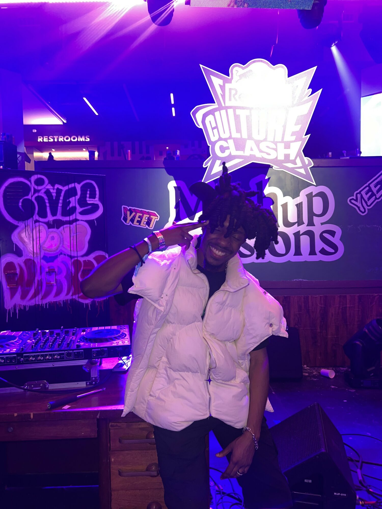 Person with dreadlocks in a white vest making a peace sign at a gaming event with bright purple lighting.