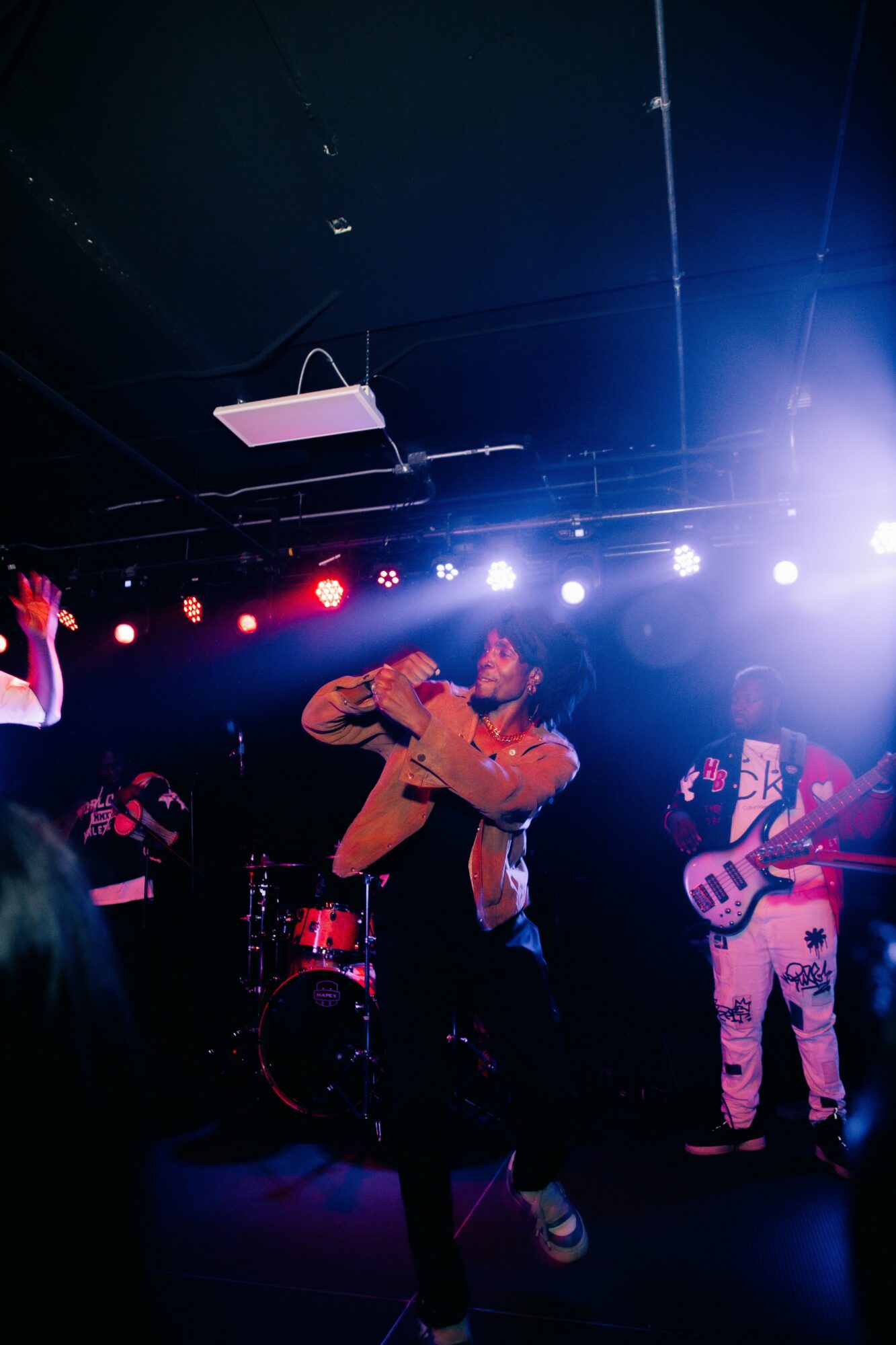 Band performing on stage with colorful lighting, one person singing into a microphone, others playing instruments, dark background.