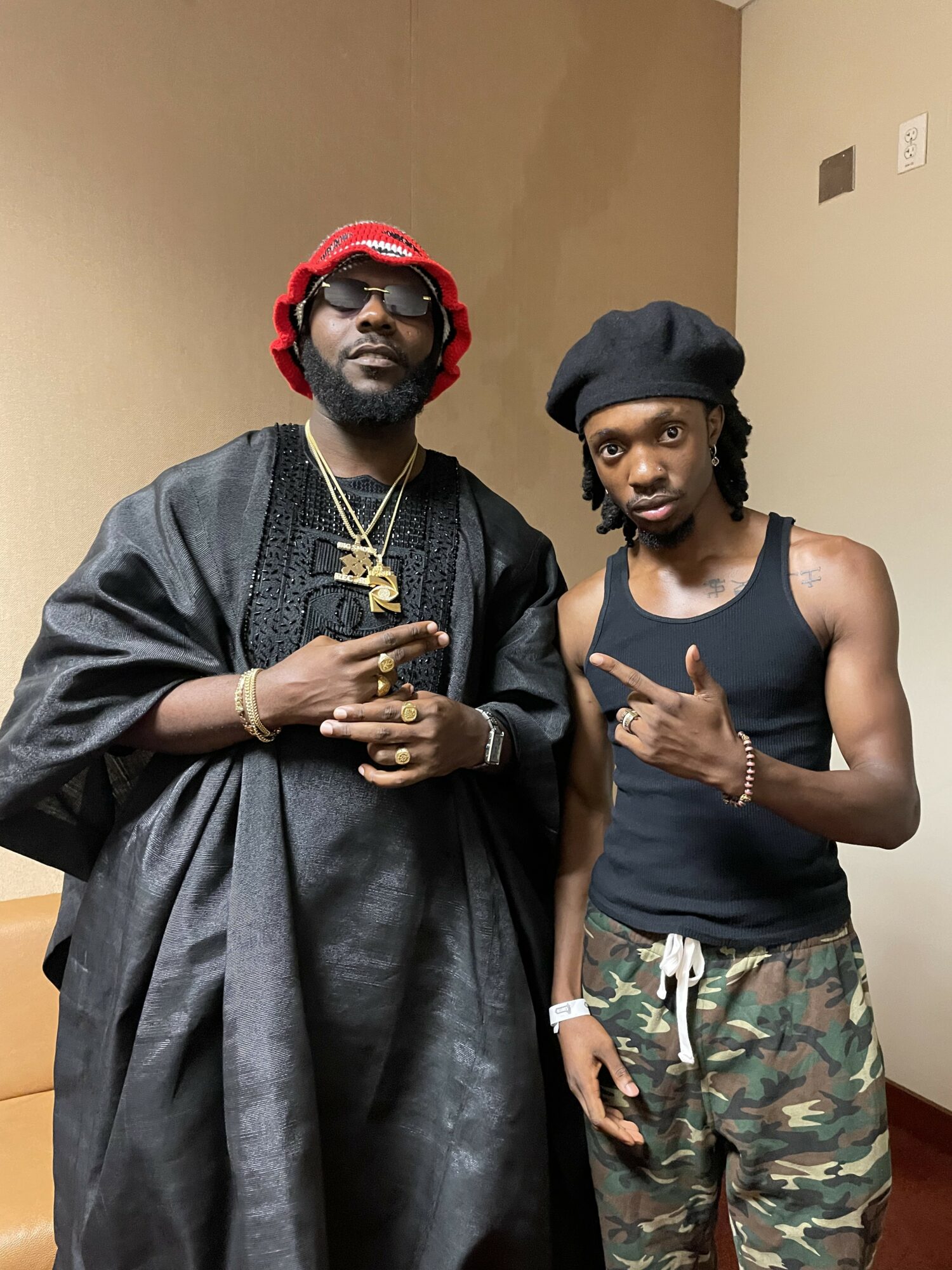 Two men standing indoors, one in traditional attire with jewelry and the other in casual clothing, pointing at each other.