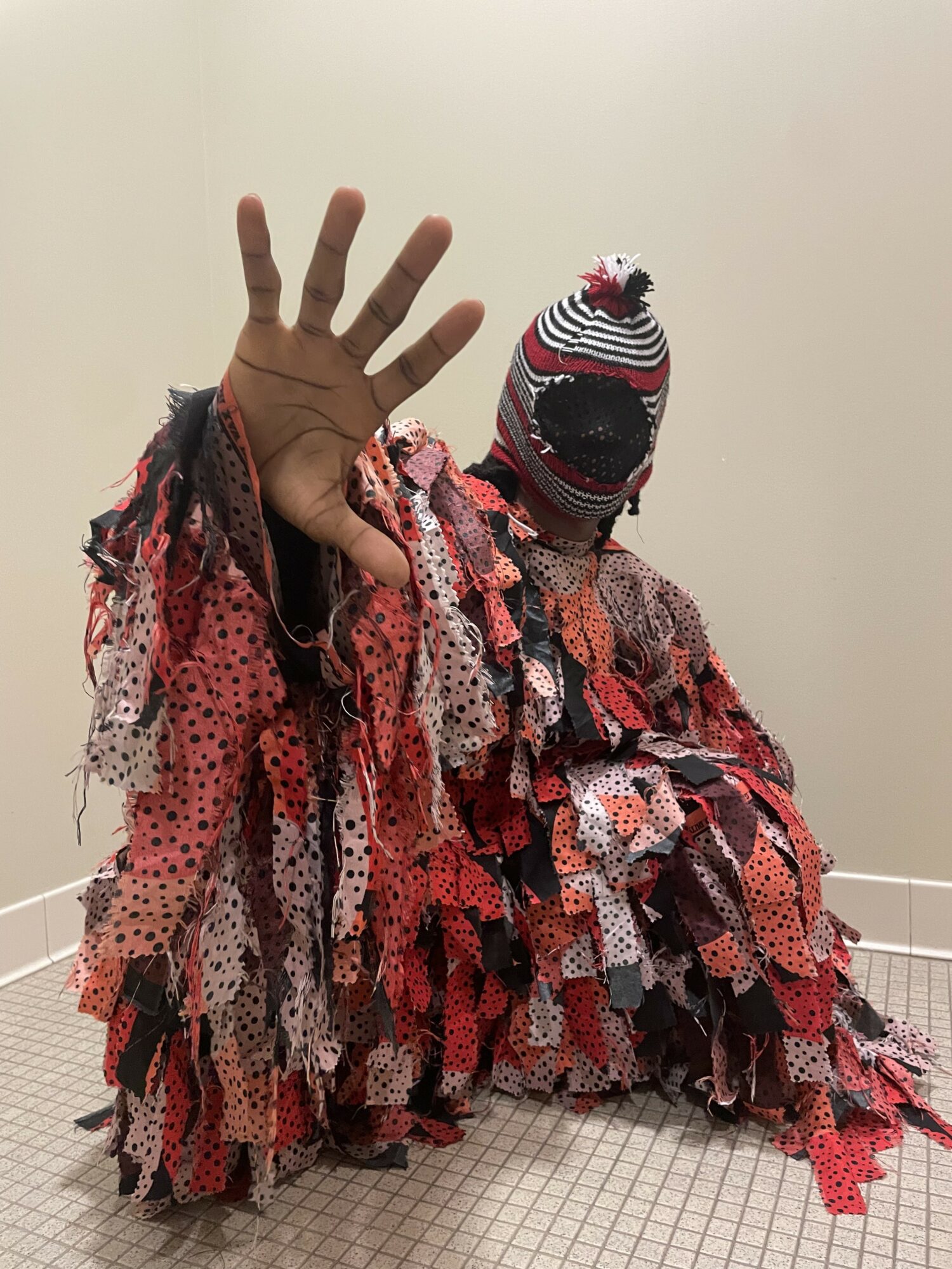 Person wearing a multicolored, layered, patchwork costume with a striped hat, raising a hand, against a plain wall.