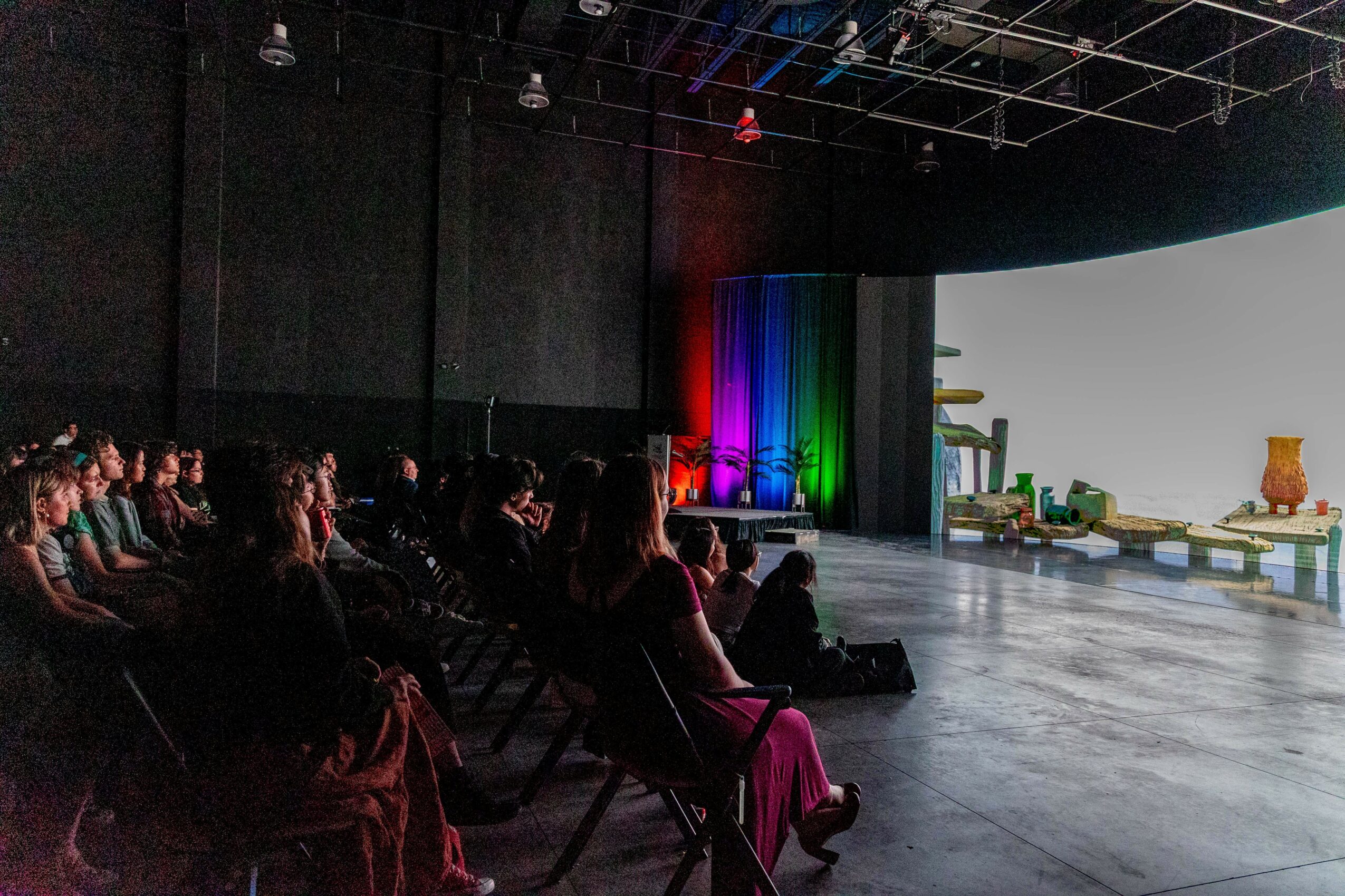 Audience seated in a dark theater watching a brightly lit stage with colorful lights and props.