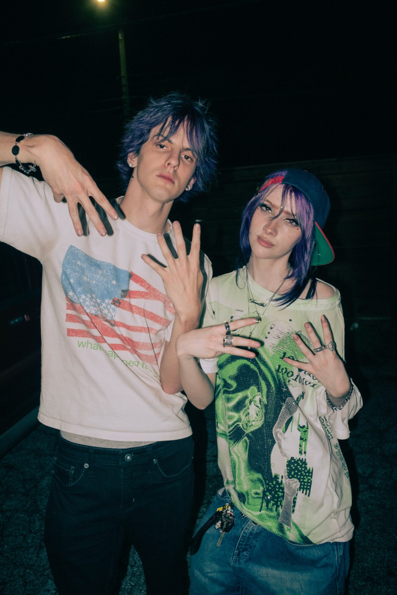 Two young people with colorful hair pose with hands raised, wearing casual clothing, in a dark setting.