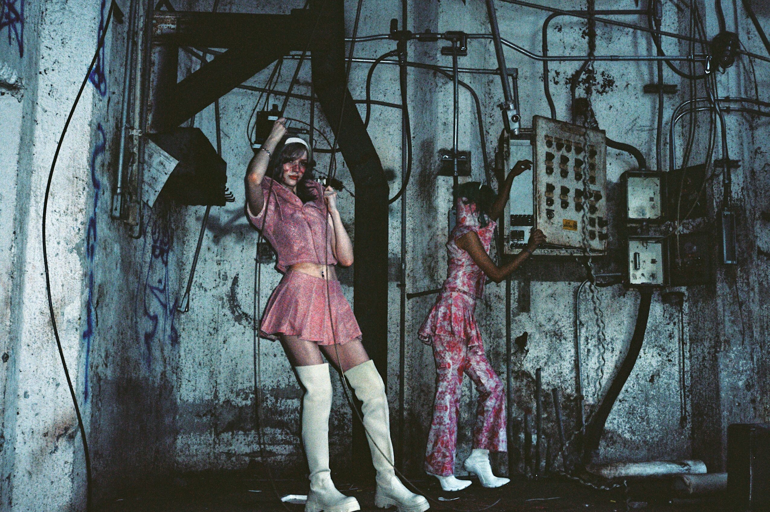 Two performers on stage with a backdrop of wires and electrical equipment, one in a pink dress and white boots, the other in patterned clothing, performing with microphones.