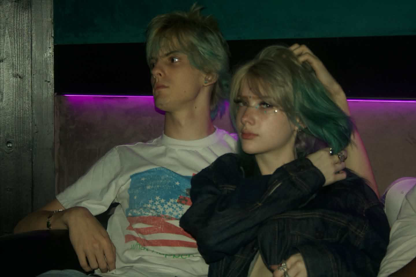 Two young people with dyed hair sit close together in a dimly lit room with purple lighting, one resting their head on the other's shoulder.