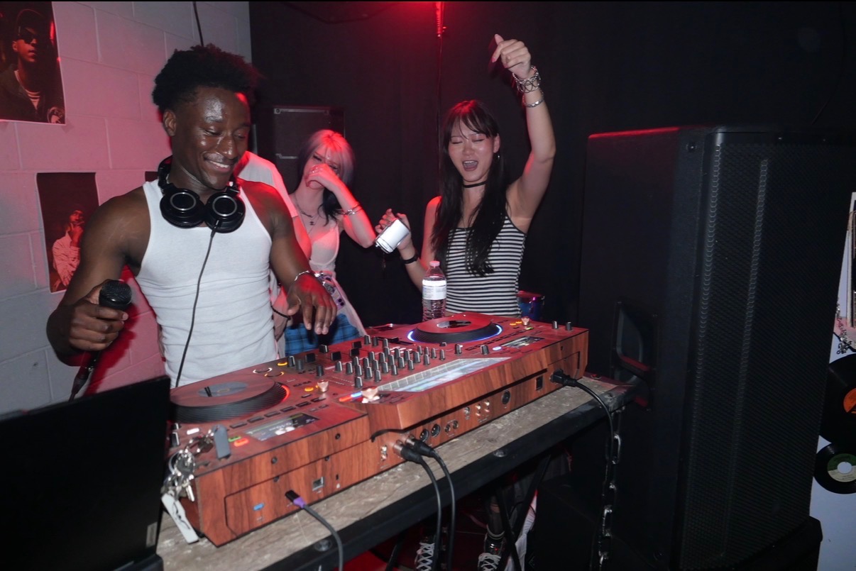 Two women dancing and DJing at a party with red lighting, one with headphones around her neck, smiling and raising her arm.