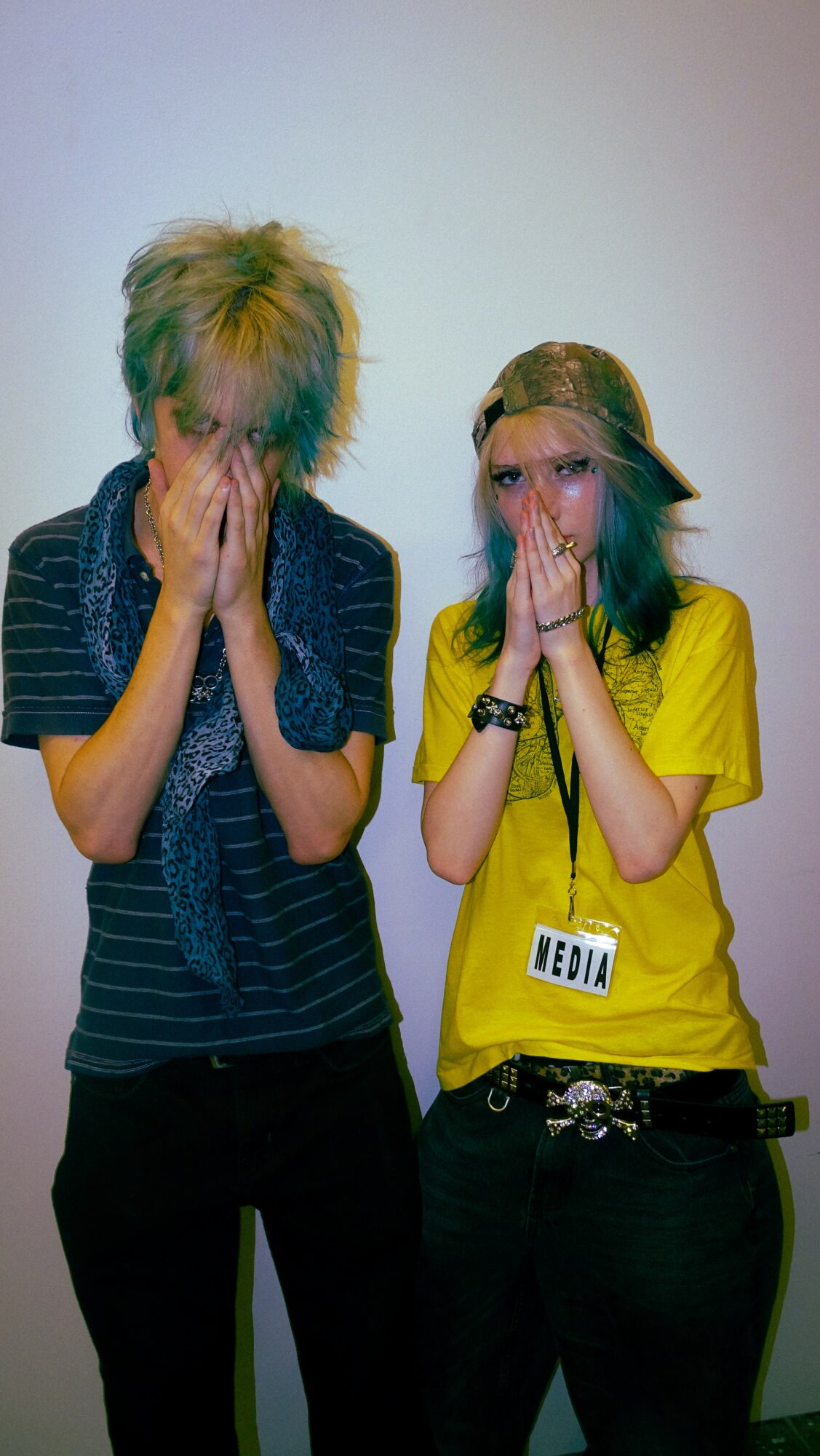 Two people with hands covering their faces, standing against a plain background.
