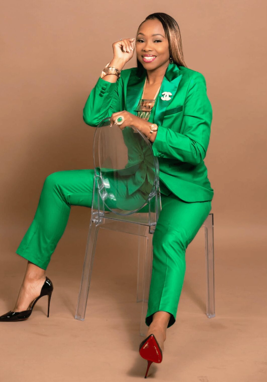Woman in green suit sitting on transparent chair, smiling, with a brown background.