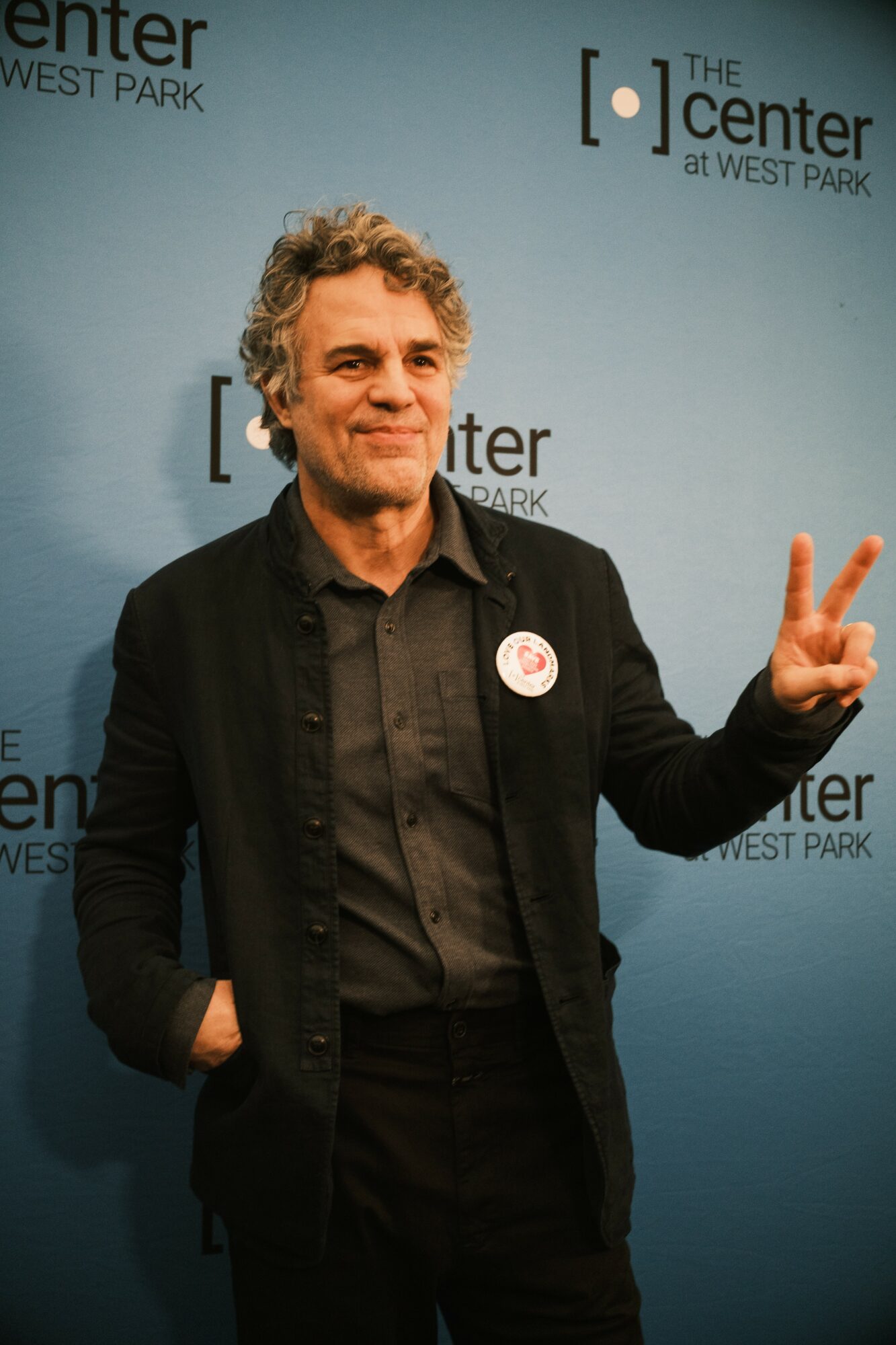 Man with curly hair wearing a black jacket and shirt, making a peace sign gesture, standing in front of a blue background with text.