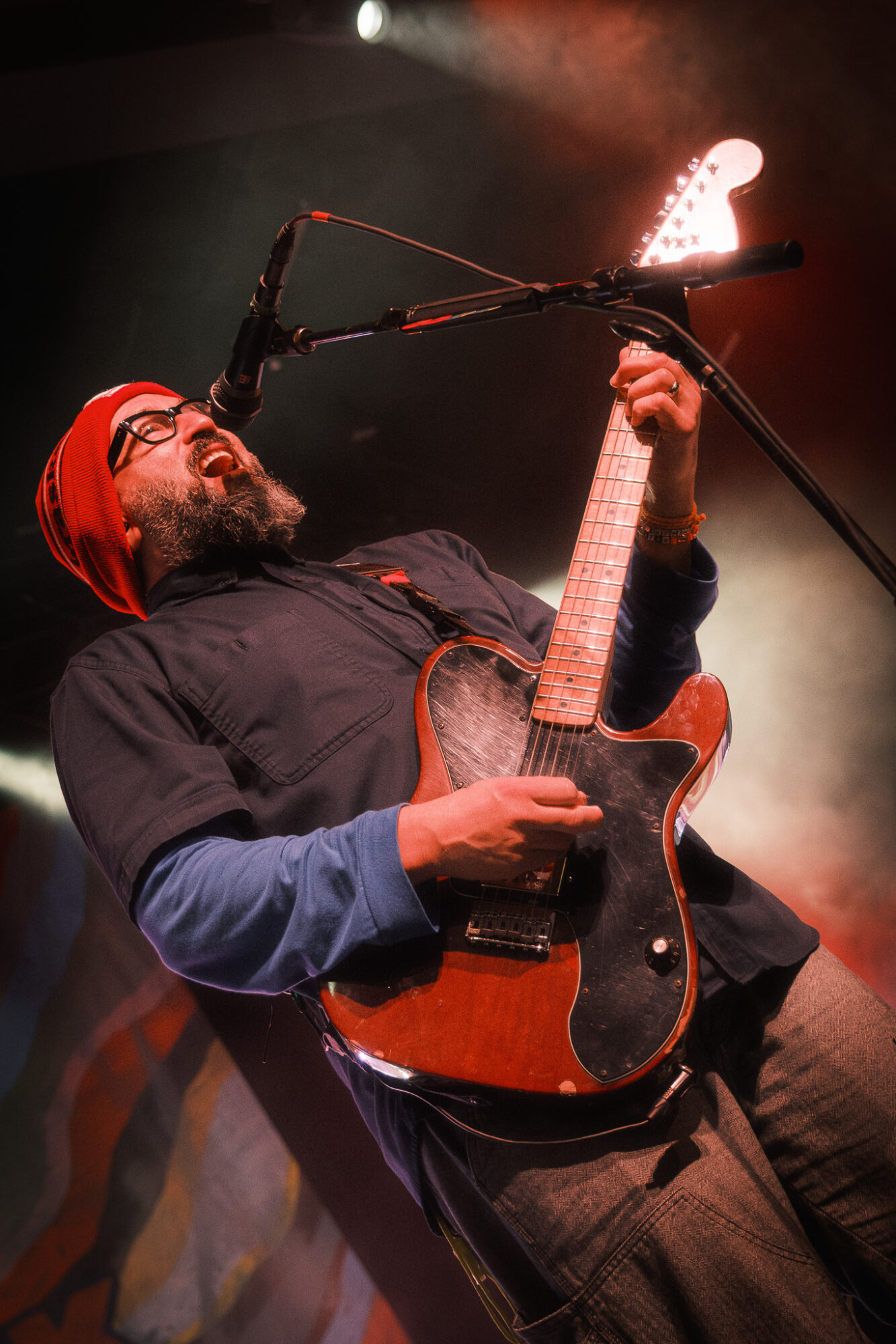 Person playing an electric guitar on stage with smoke and lighting effects, wearing glasses and a red head covering.