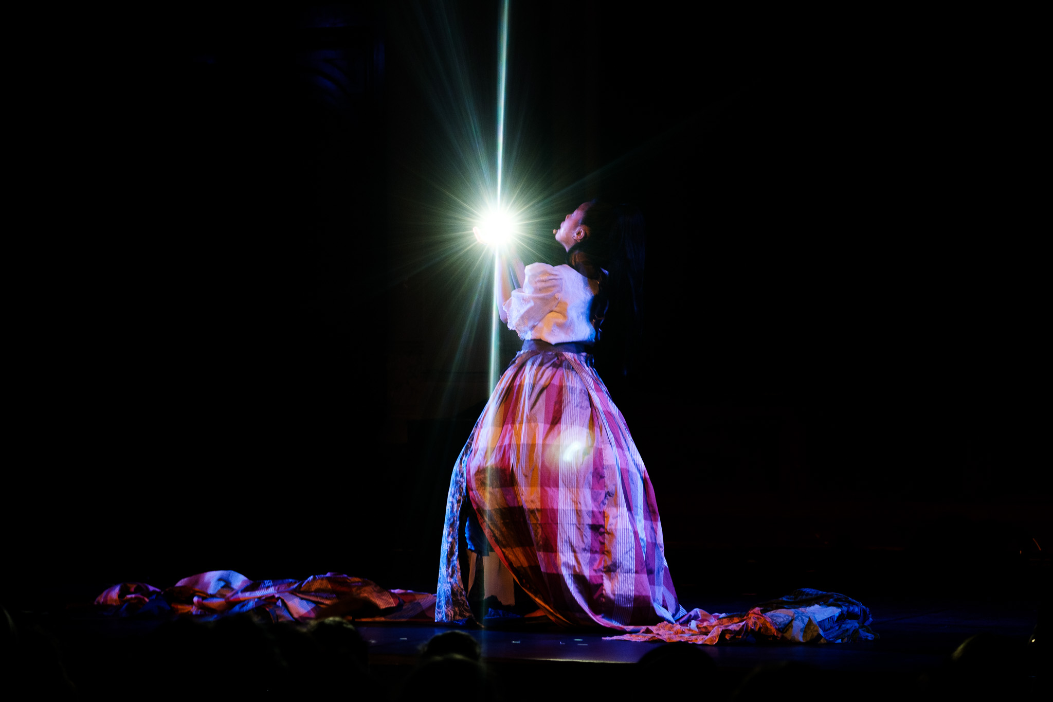Person standing on stage with a bright light behind, wearing a long, colorful skirt and a white top.