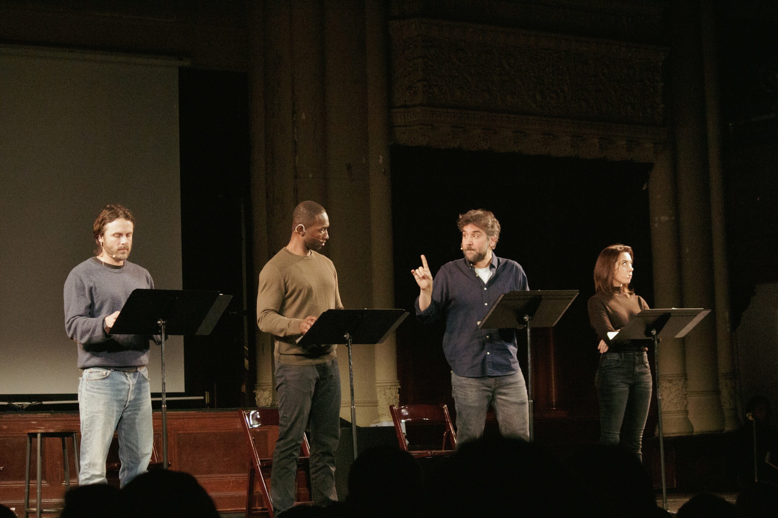 Four people stand on stage with music stands, one person appears to be conducting or speaking, in a theater setting.