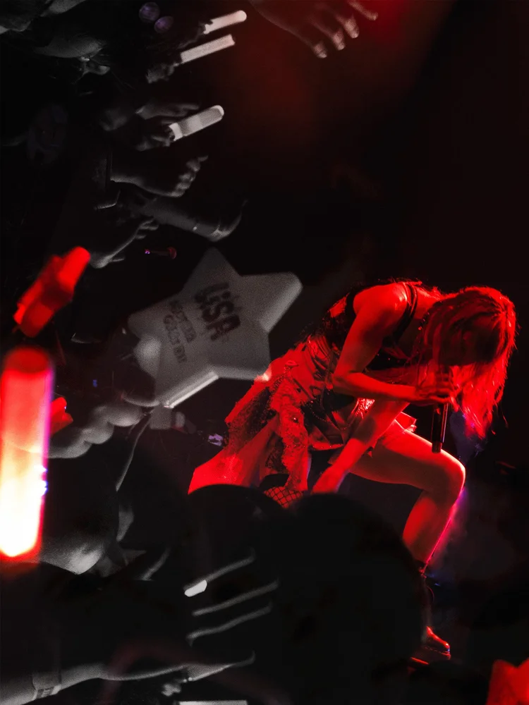 Performer with long hair on stage, illuminated by red light, holding a microphone, with stage equipment visible in background.