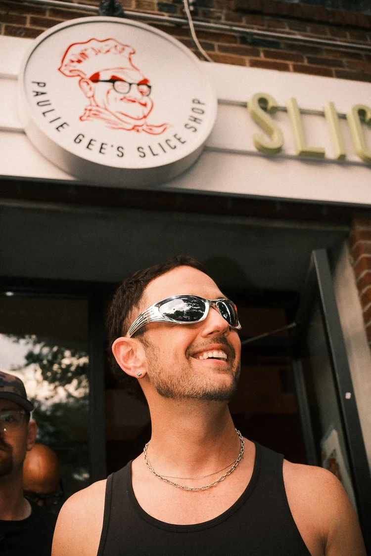Smiling man wearing sunglasses and a black tank top outside a shop with a sign featuring a cartoon face and text.