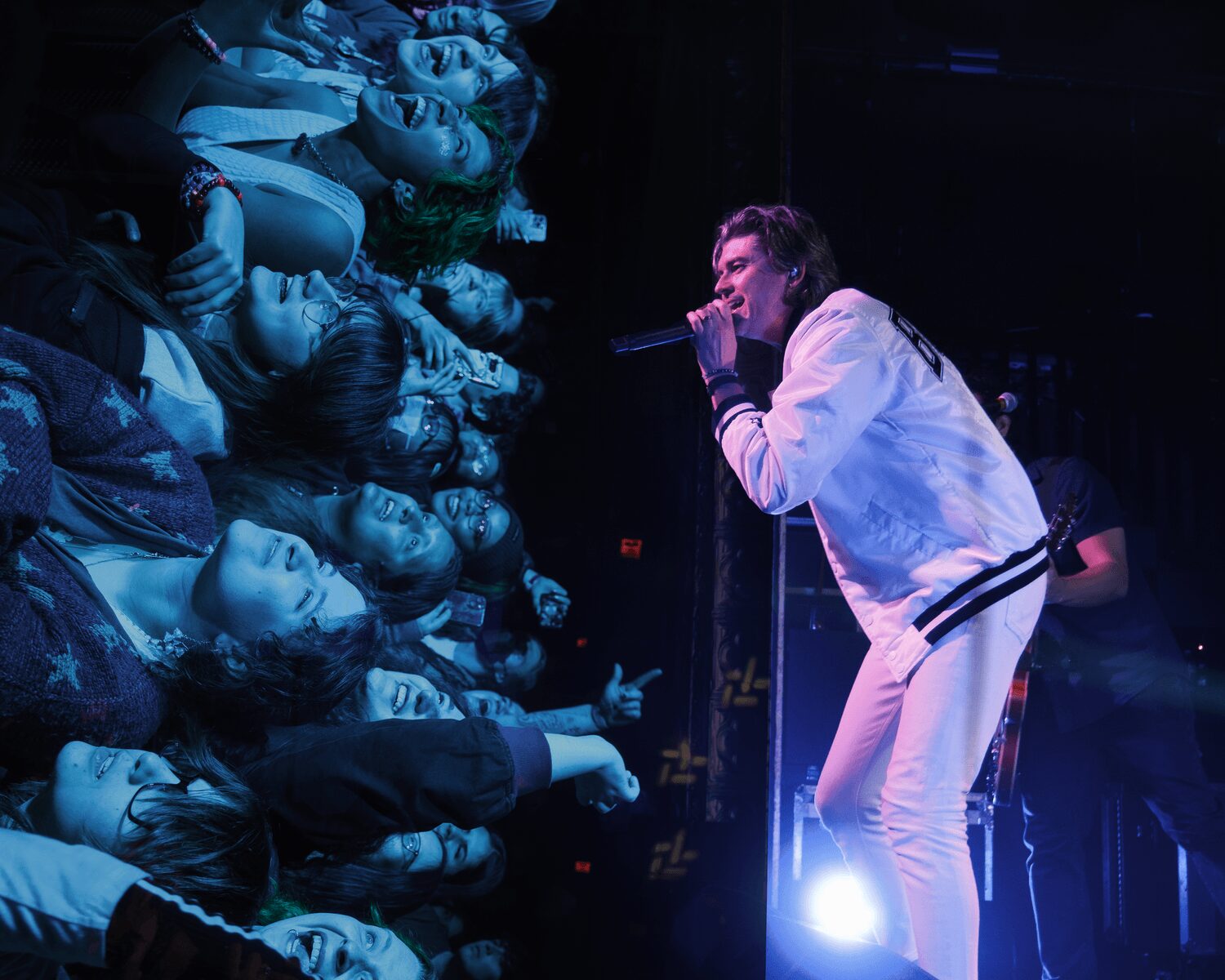 Performer singing into microphone on stage with audience reaching out, blue lighting, and dark background.