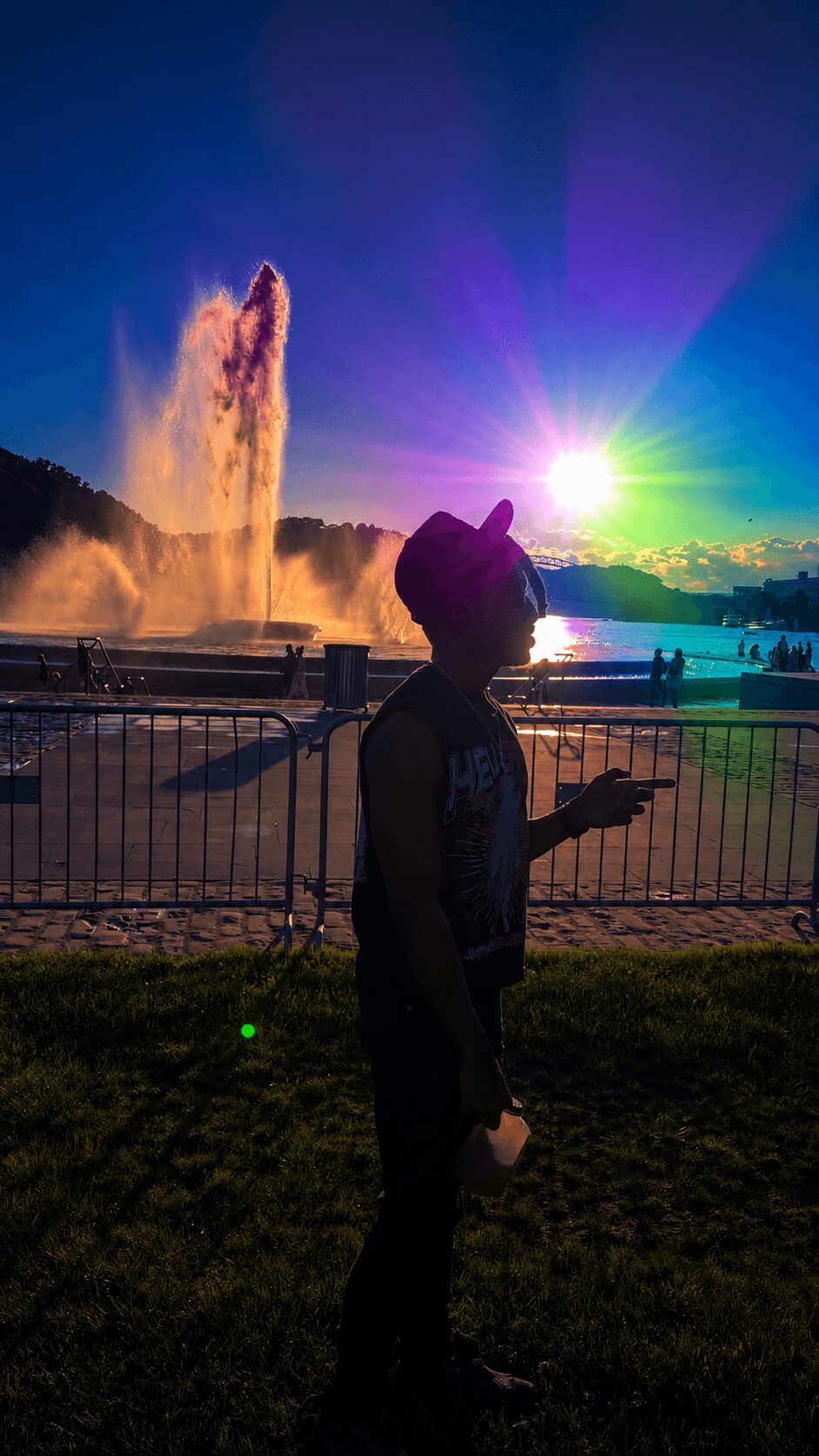 Person wearing a hat standing near a fence, with a colorful sunset and water fountain in the background.