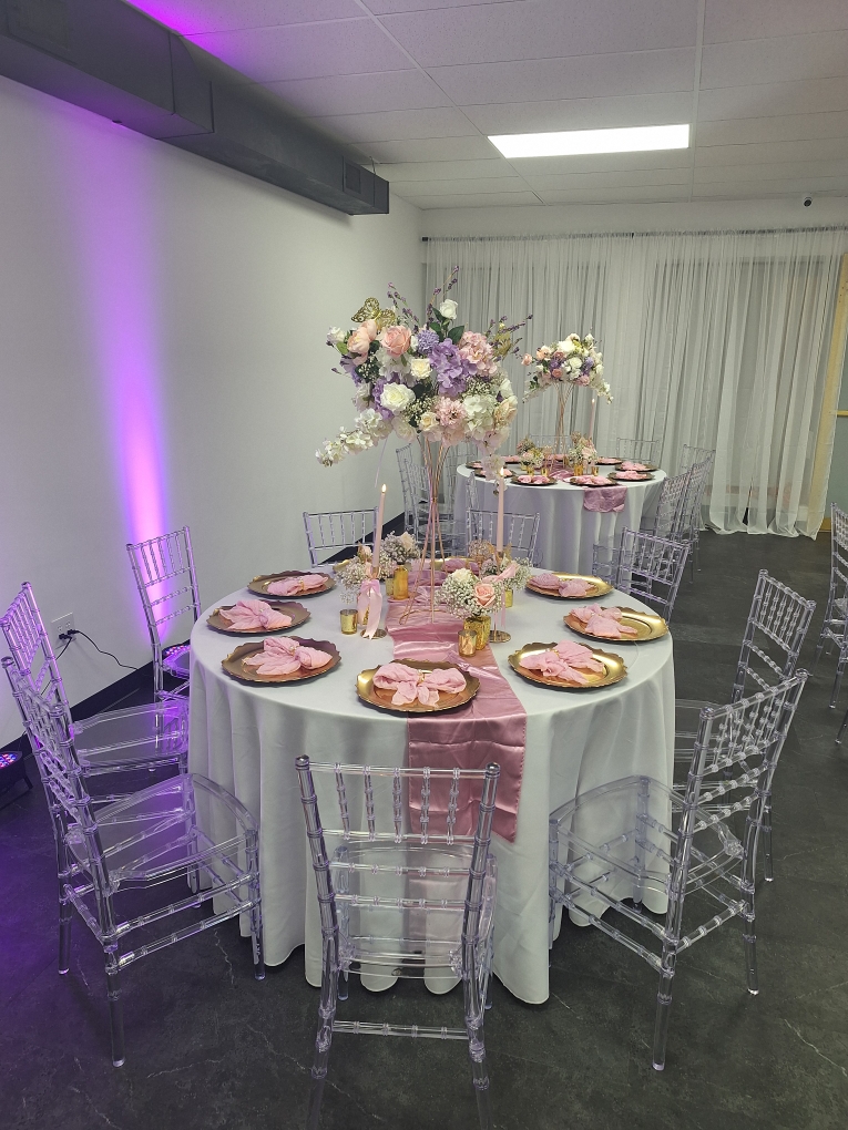 Round table with floral centerpiece and pink napkins, surrounded by clear chairs, in a decorated event space.