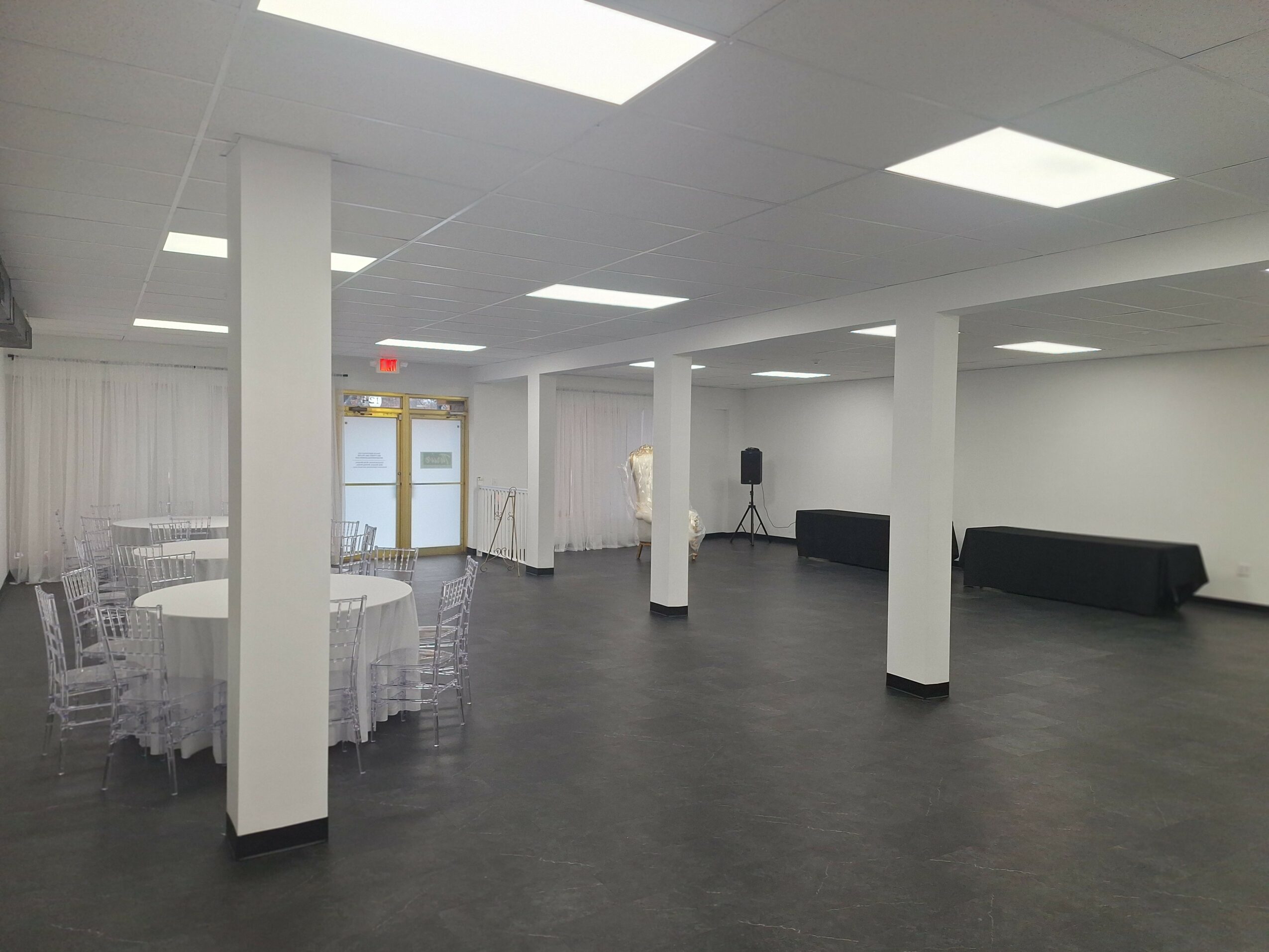 Empty event space with round tables, chairs, white walls, and ceiling lights, with a door and window in the background.