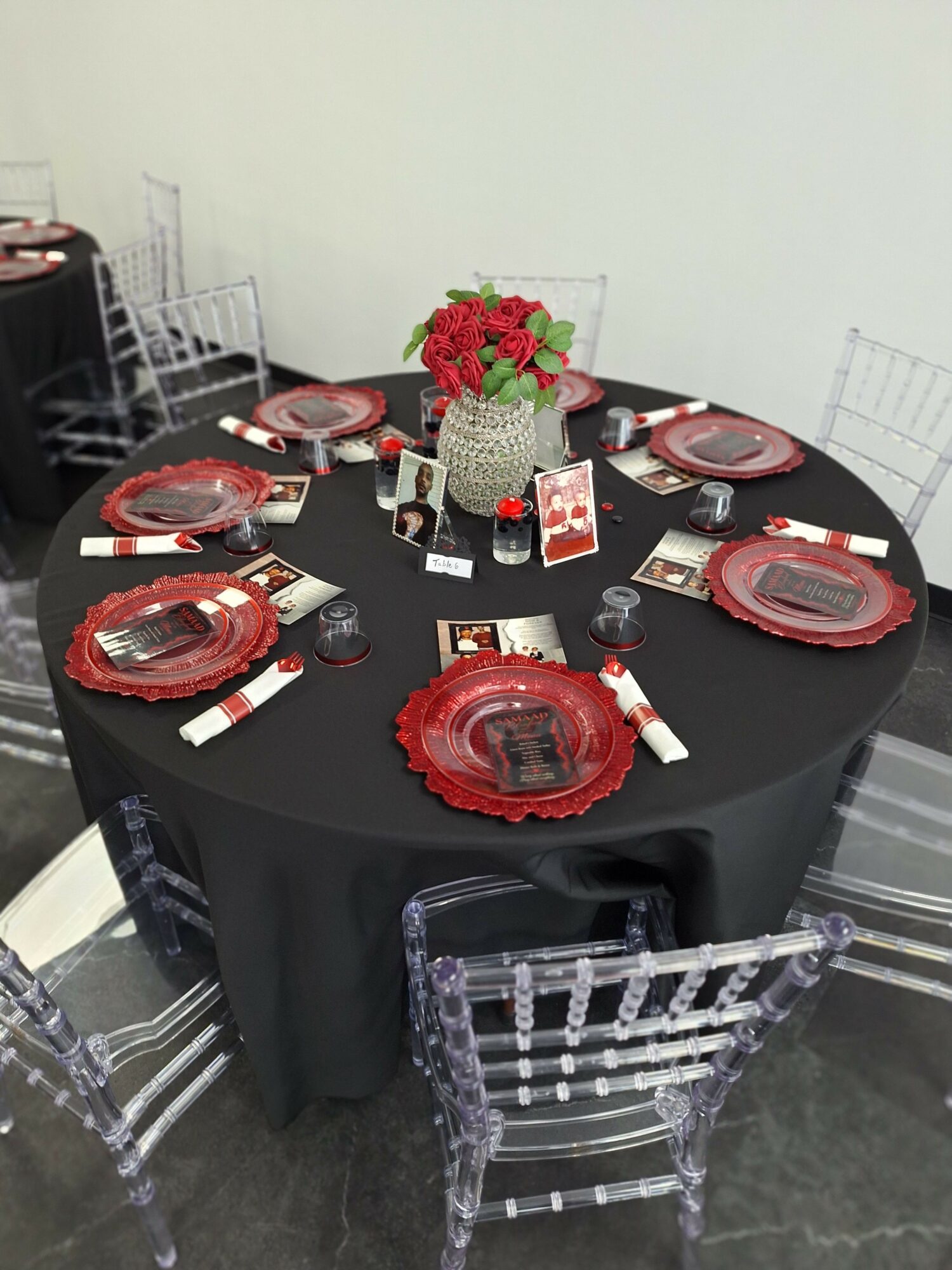 Round table set with red plates, glasses, cutlery, a flower centerpiece, and small decorative items, with transparent chairs around it.