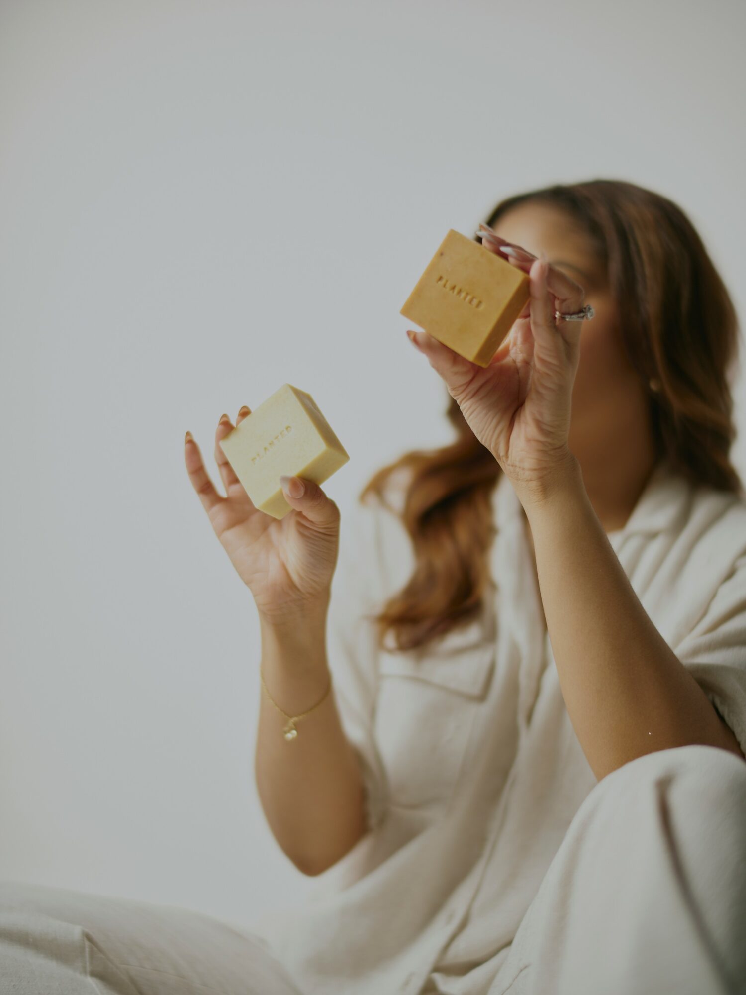 Woman with long hair holding two small boxes, one near her face and the other in her hand, against a plain background.