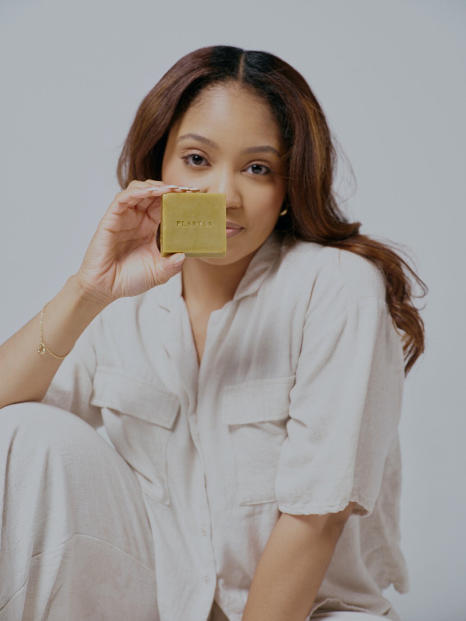 Woman with long hair holding a yellow sponge near her face, wearing a light-colored shirt, against a plain background.