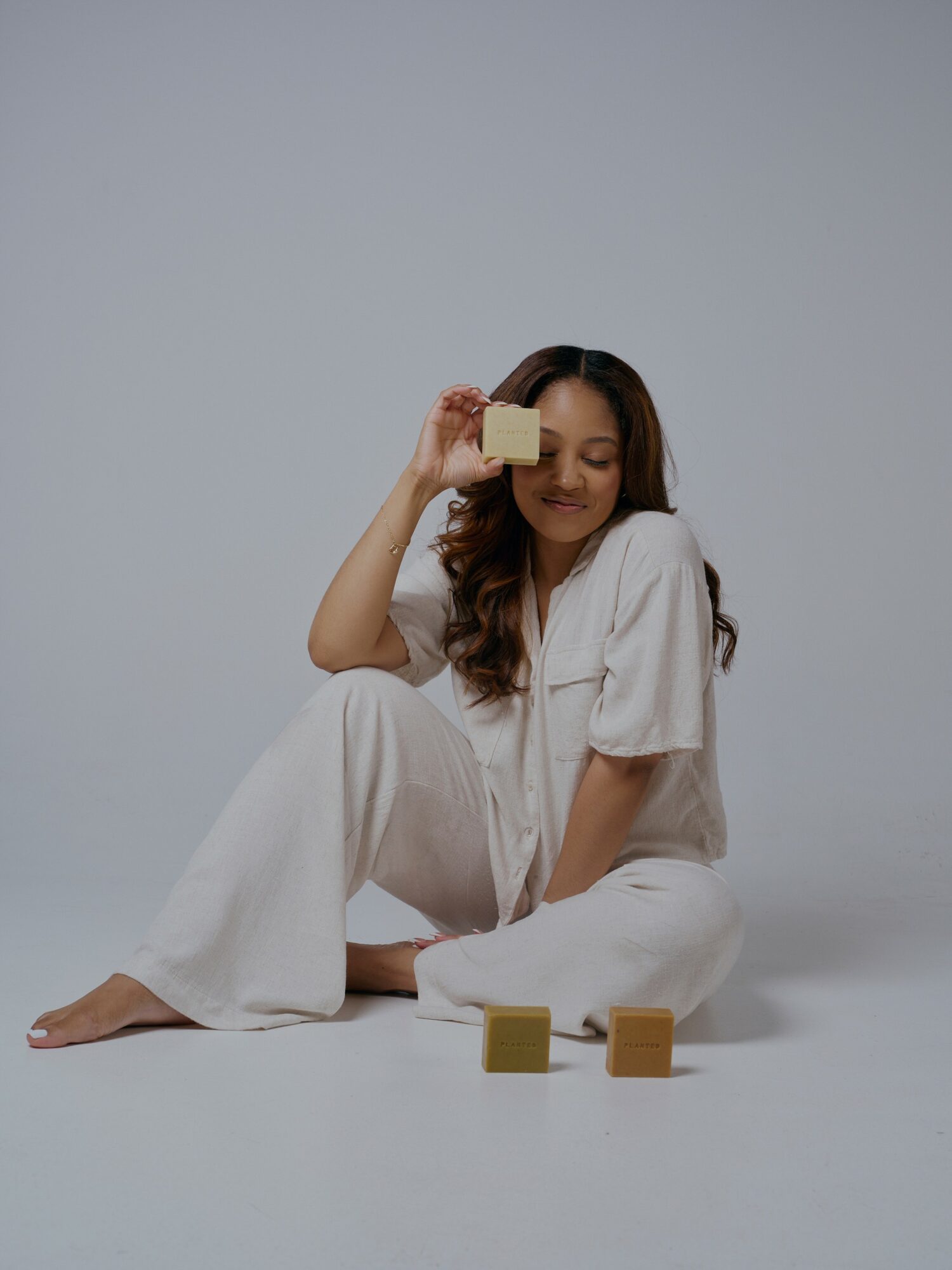 Woman sitting on the floor holding a soap bar to her eye, with soap bars in front, against a plain background.