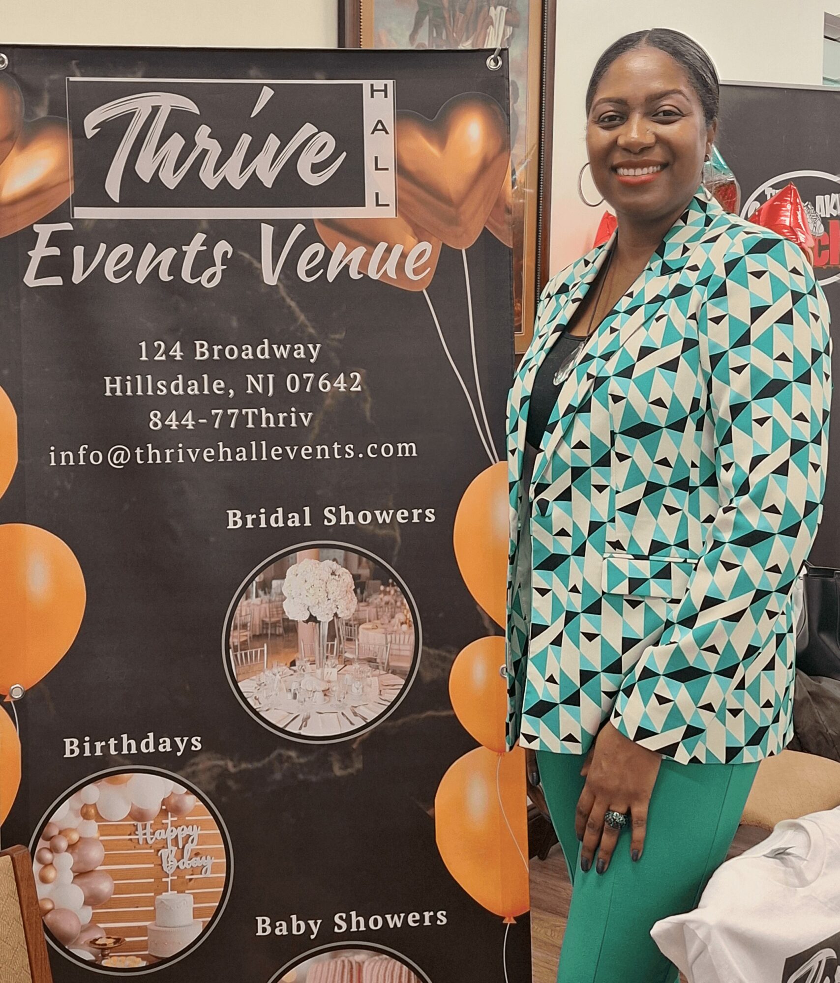 Smiling woman in patterned blazer and green pants standing next to event venue sign with balloons and event details.