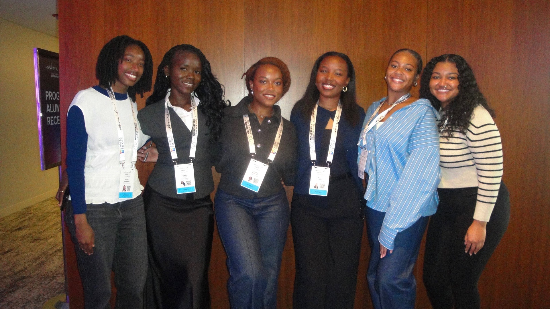 Six women standing together indoors, smiling, wearing name tags, in front of a wooden wall.
