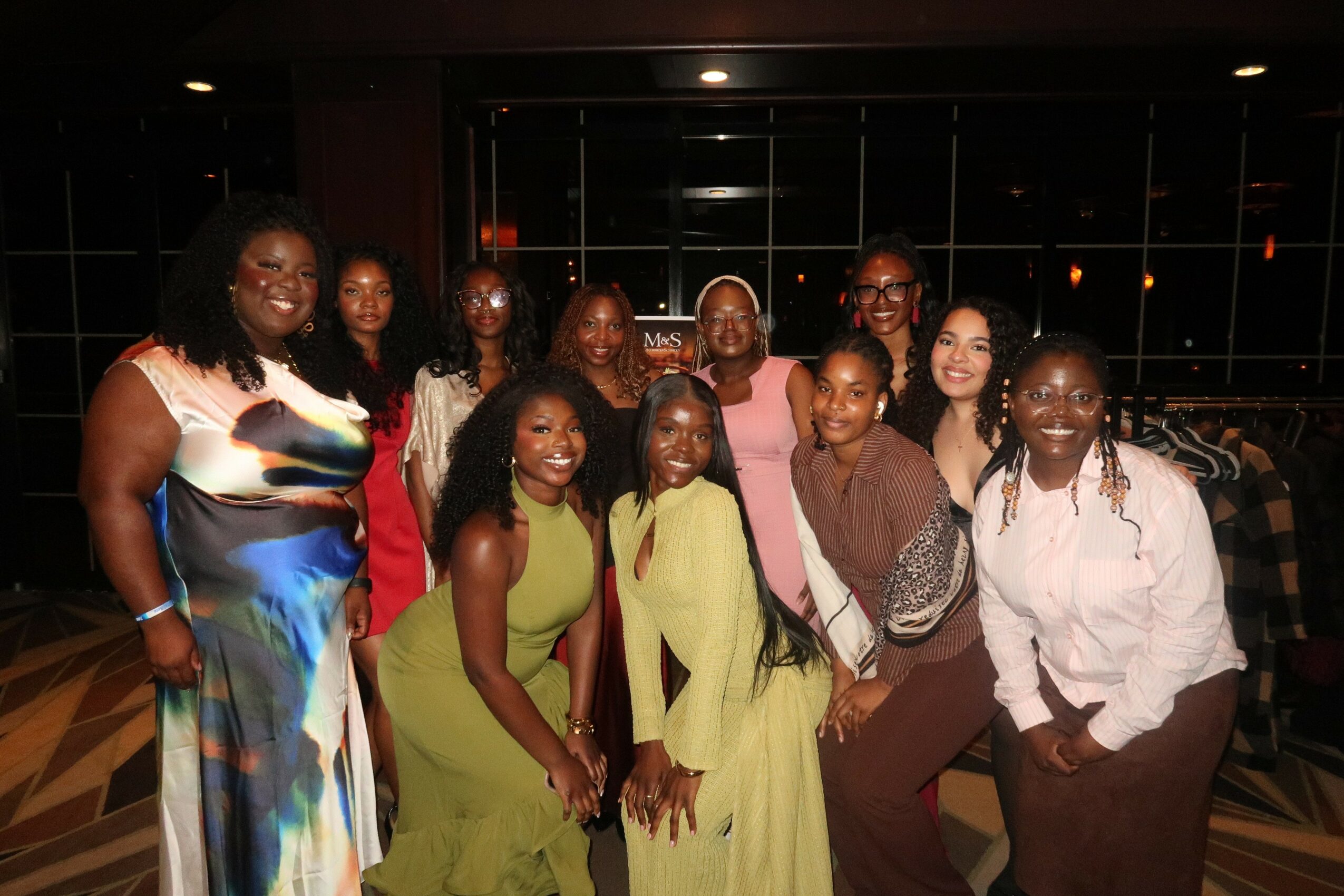 Group of ten women posing indoors with large windows in the background, some smiling and wearing colorful clothing.