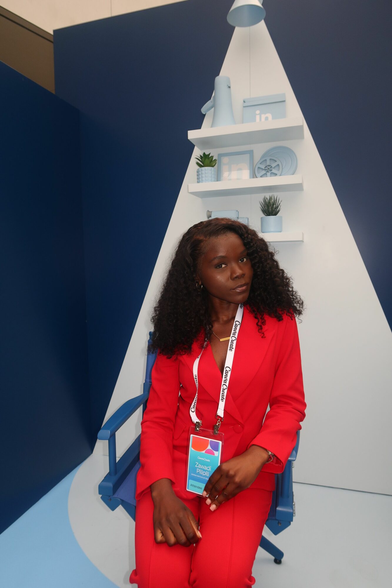 Woman with curly hair in red outfit holding a phone and badge, standing in front of a white and blue structure with shelves.