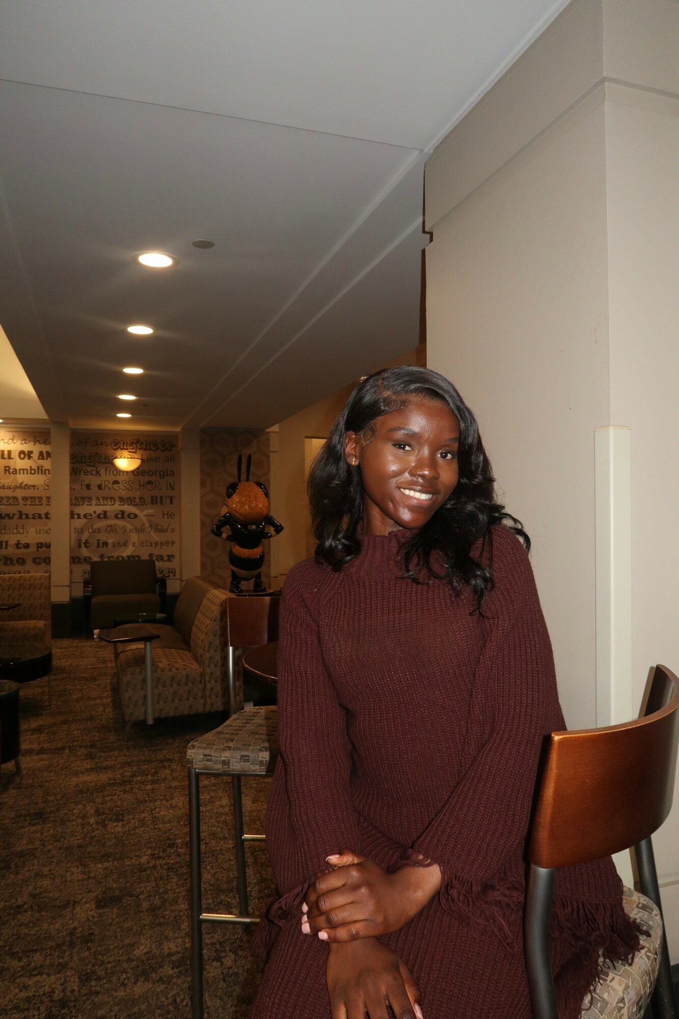 Young woman with long dark hair smiling, wearing a maroon sweater, standing indoors near a chair.