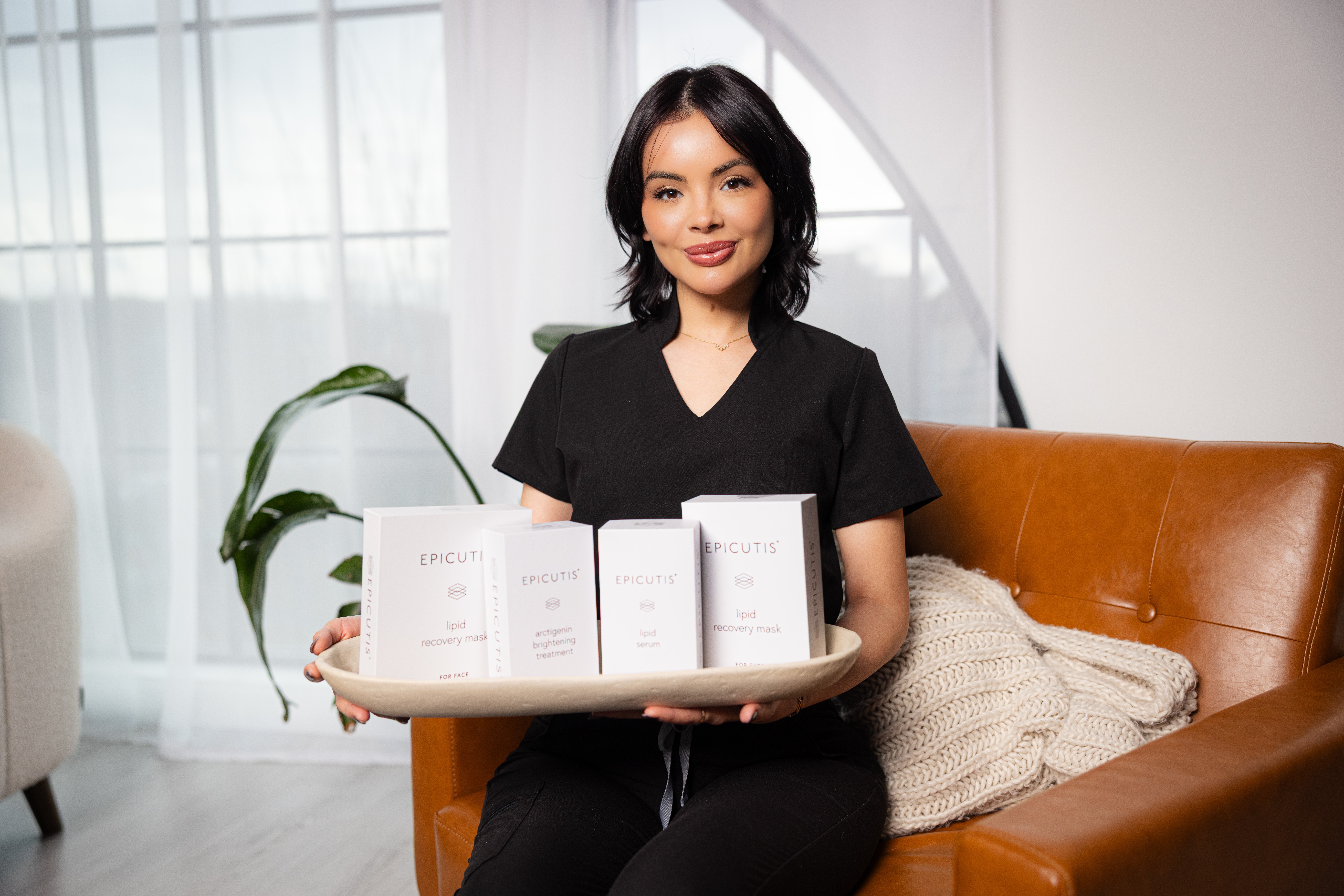 Woman in black scrubs sitting on a brown sofa holding a tray with skincare products, in a bright room with a plant and white curtains.