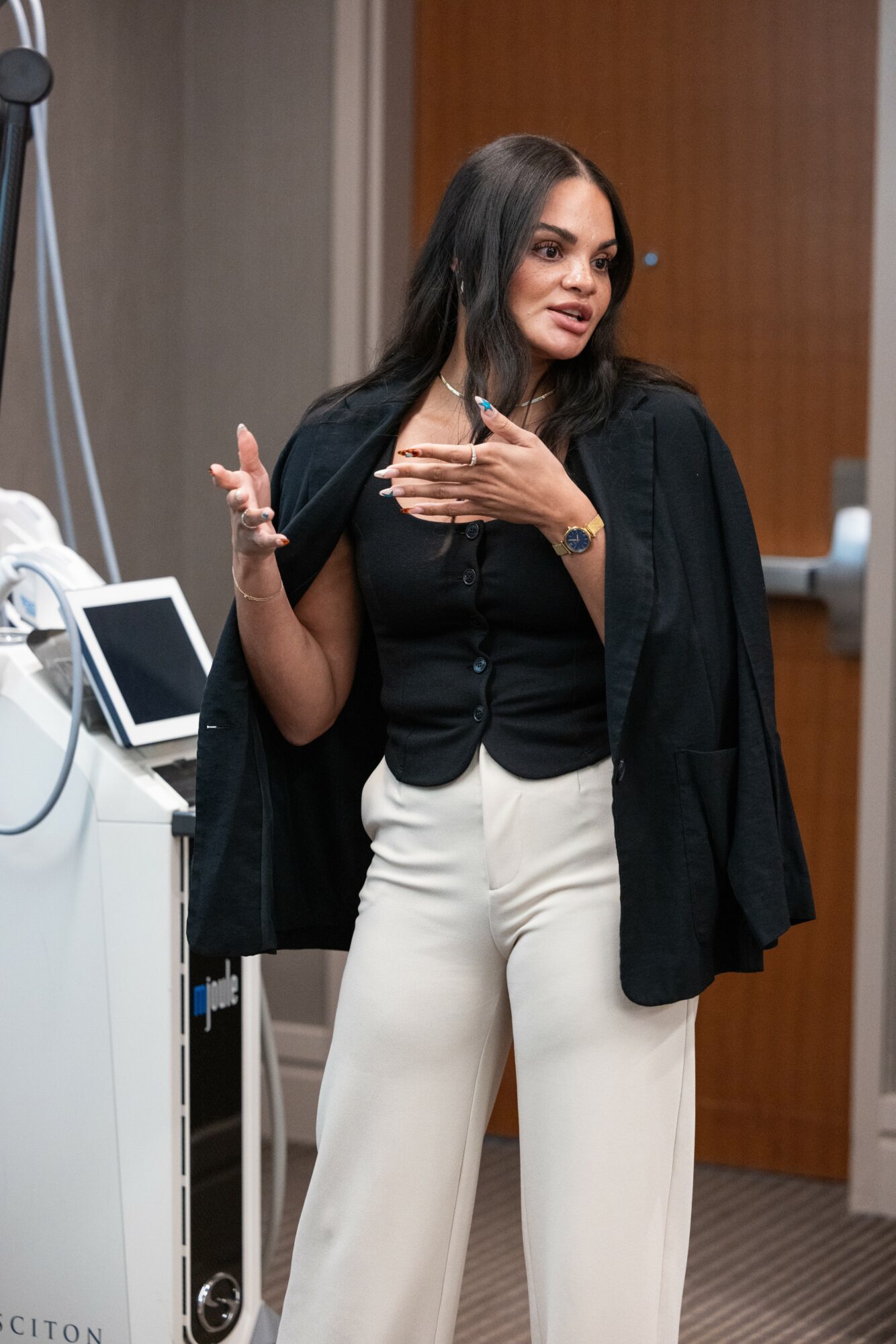 Woman with long dark hair wearing a black blazer over a black top and white pants, standing in an office.