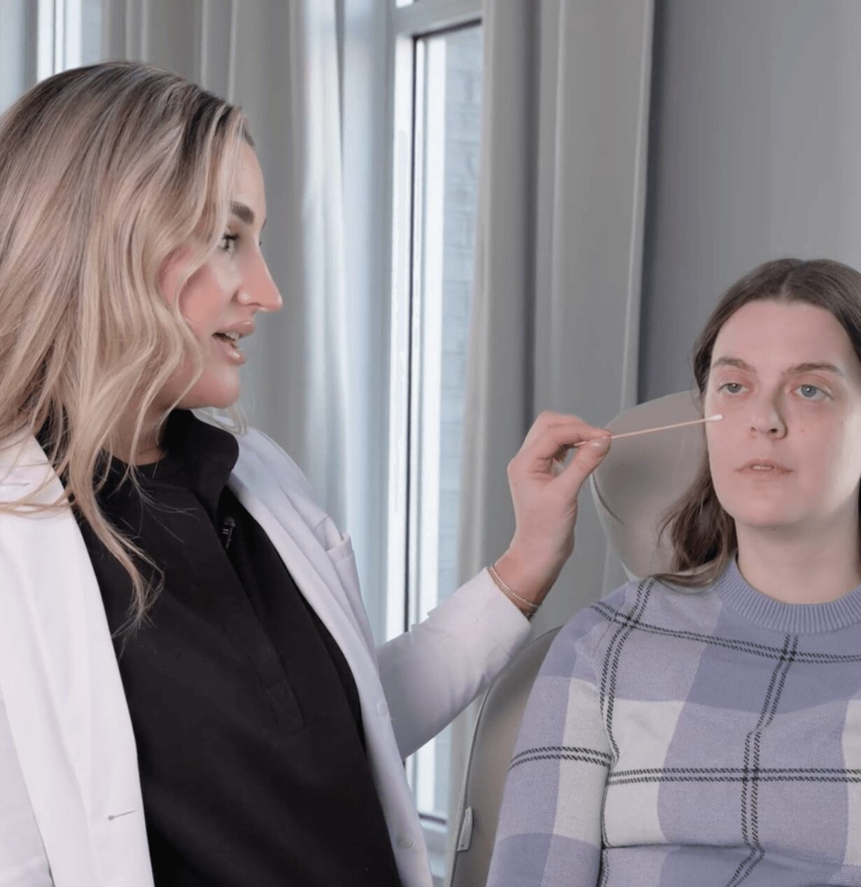 A woman applies makeup to a young girl with long hair, sitting near a window with curtains. The girl wears a plaid shirt.