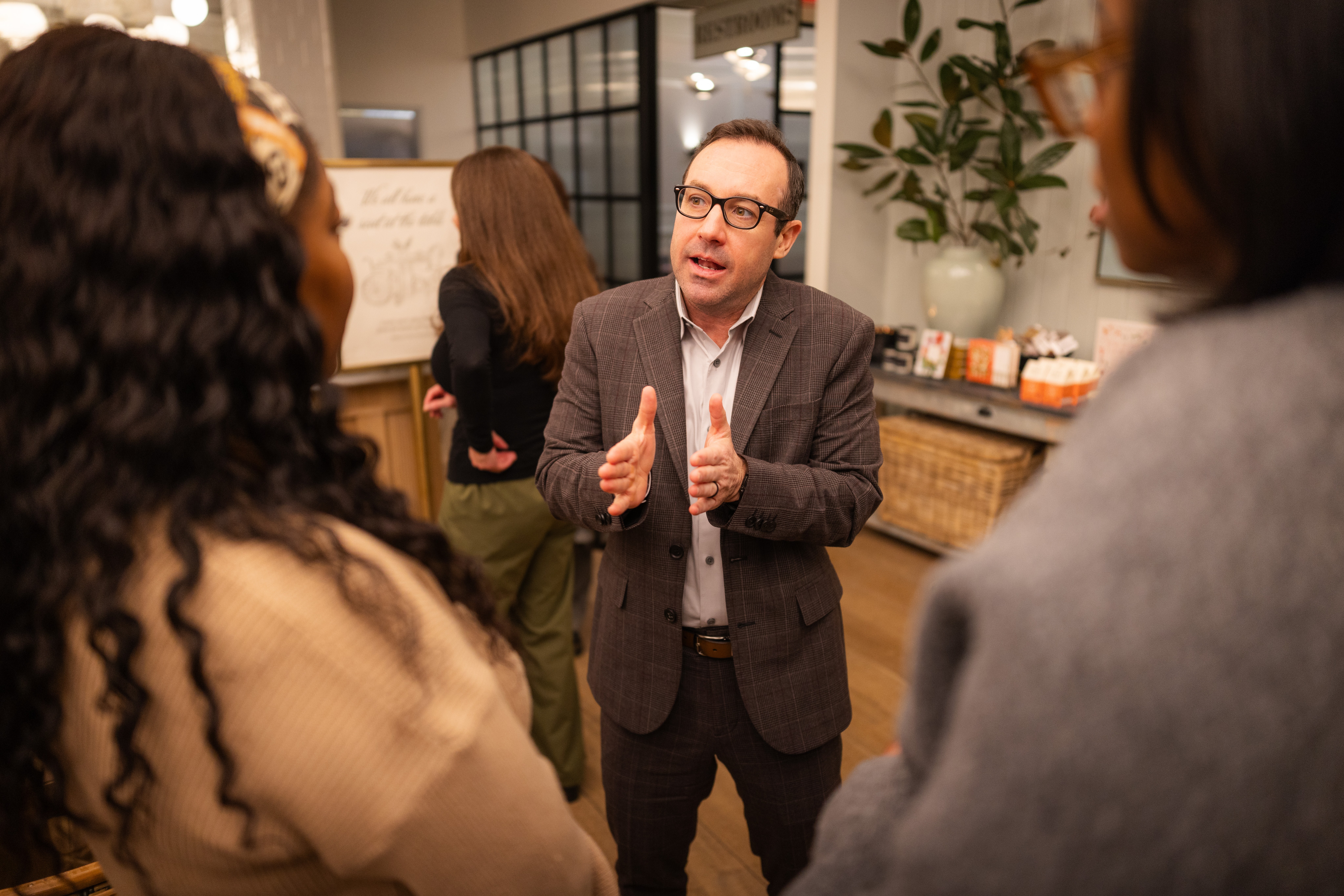 Man speaking to a group of people in an indoor setting, gesturing with hands, with plants and a whiteboard in background.