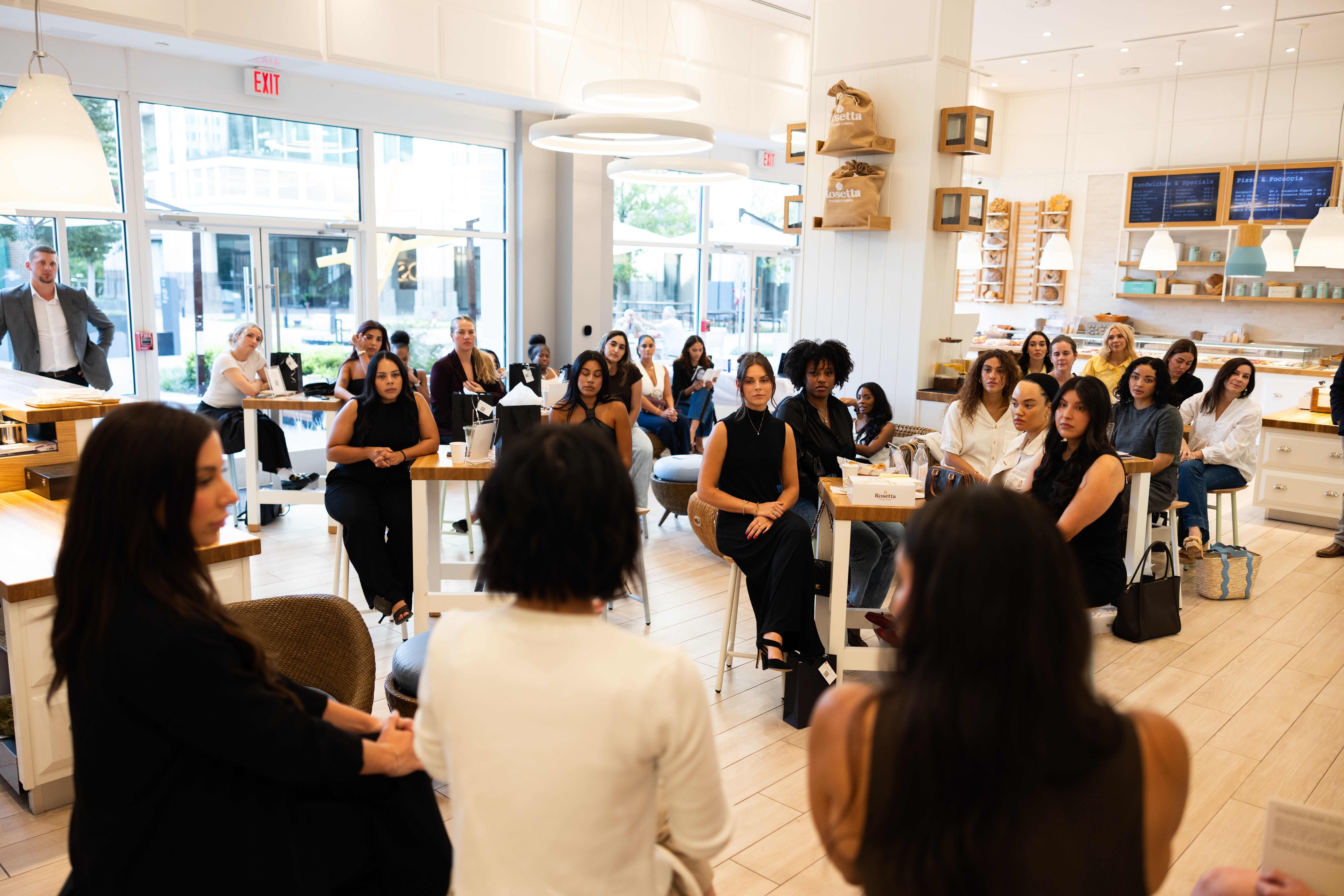 Group of people gathered in a bright, modern space with shelves and large windows, some seated and some standing.